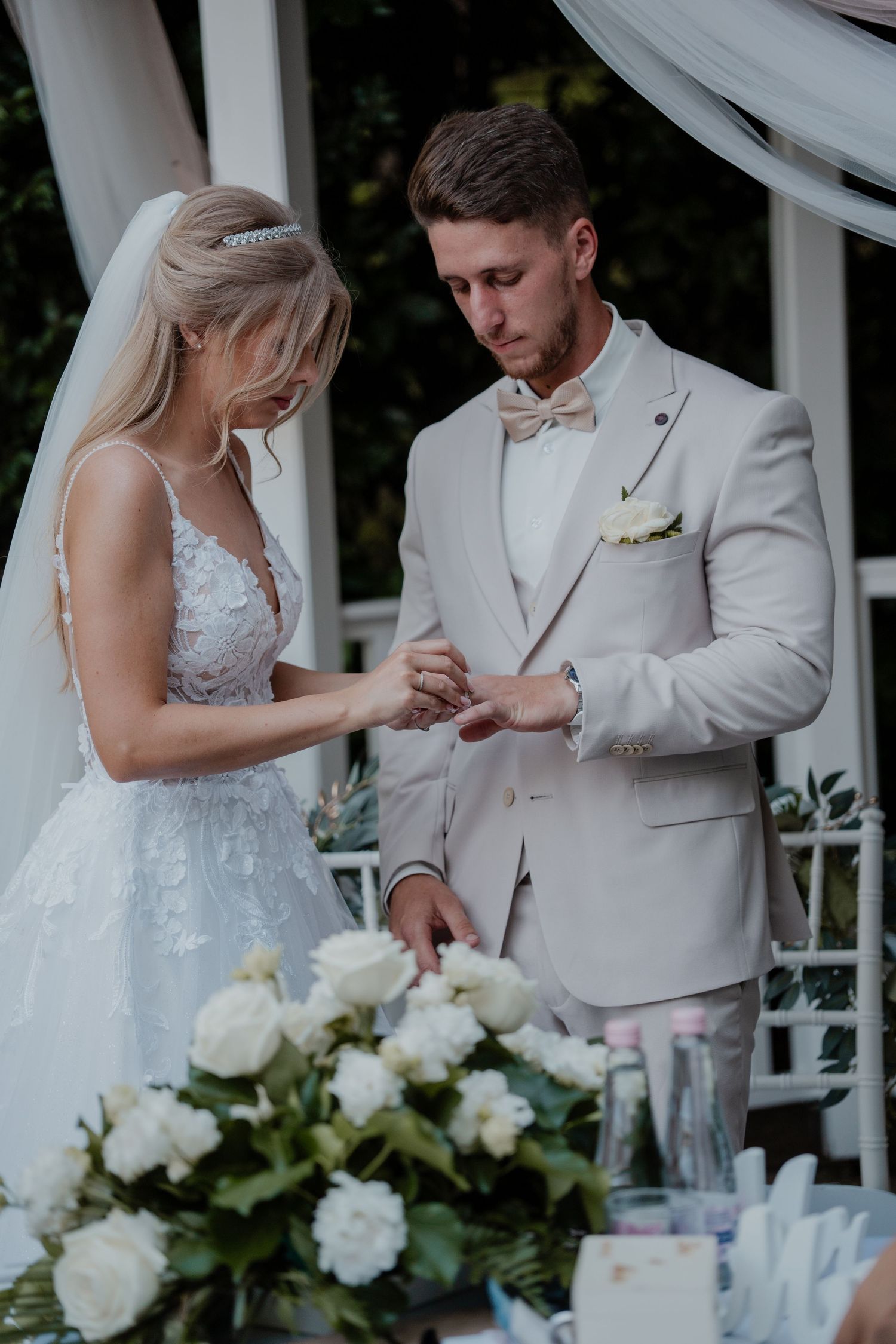 Bride placing wedding ring on groom's finger during outdoor ceremony, both dressed in elegant white and beige attire.