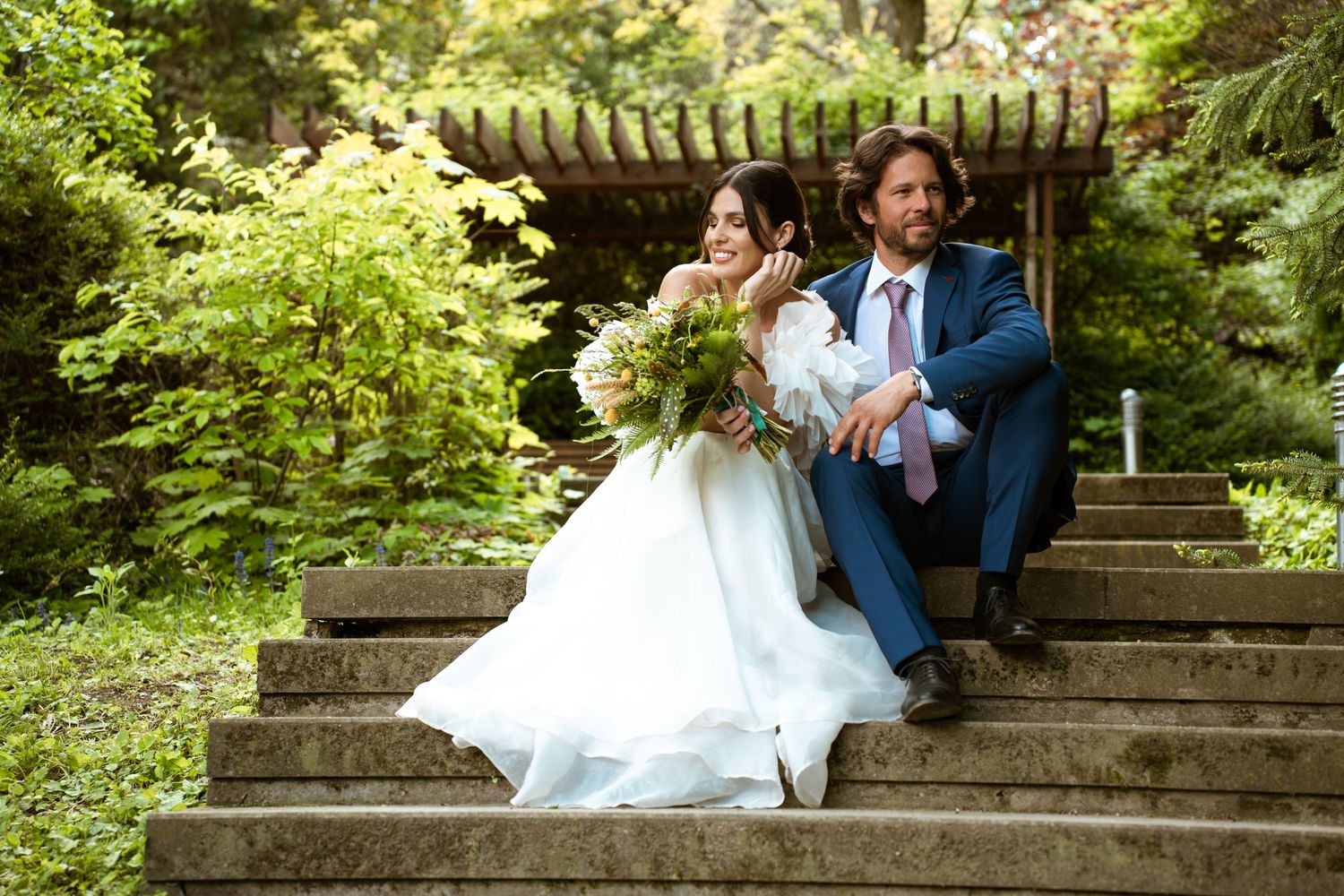 A bride and groom share a joyful moment seated on stone steps surrounded by lush garden greenery.