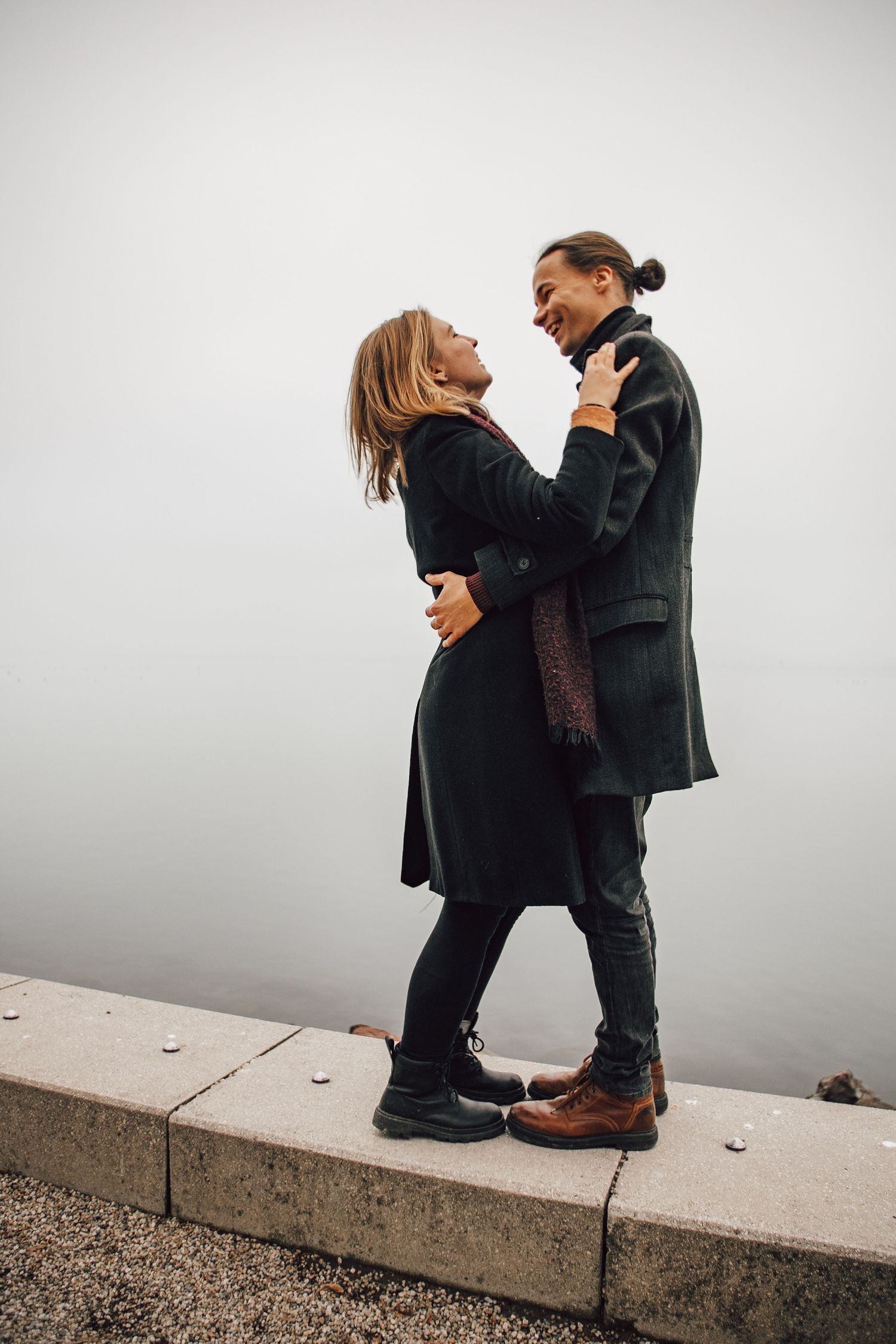 A romantic couple in dark winter coats embrace while standing on a concrete ledge against a misty gray background.