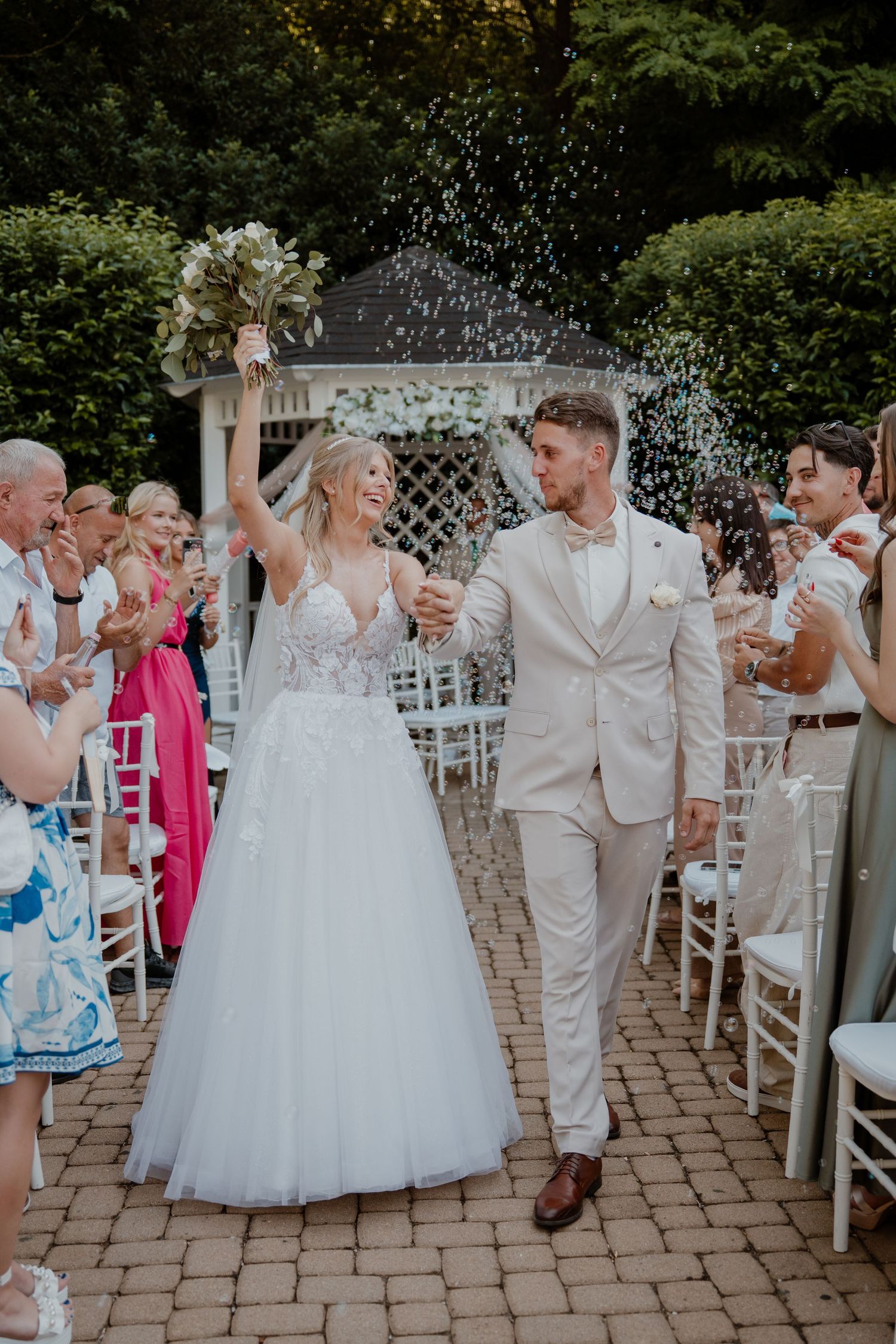 Bride and groom walk down aisle as guests toss confetti at outdoor wedding ceremony with white chairs.