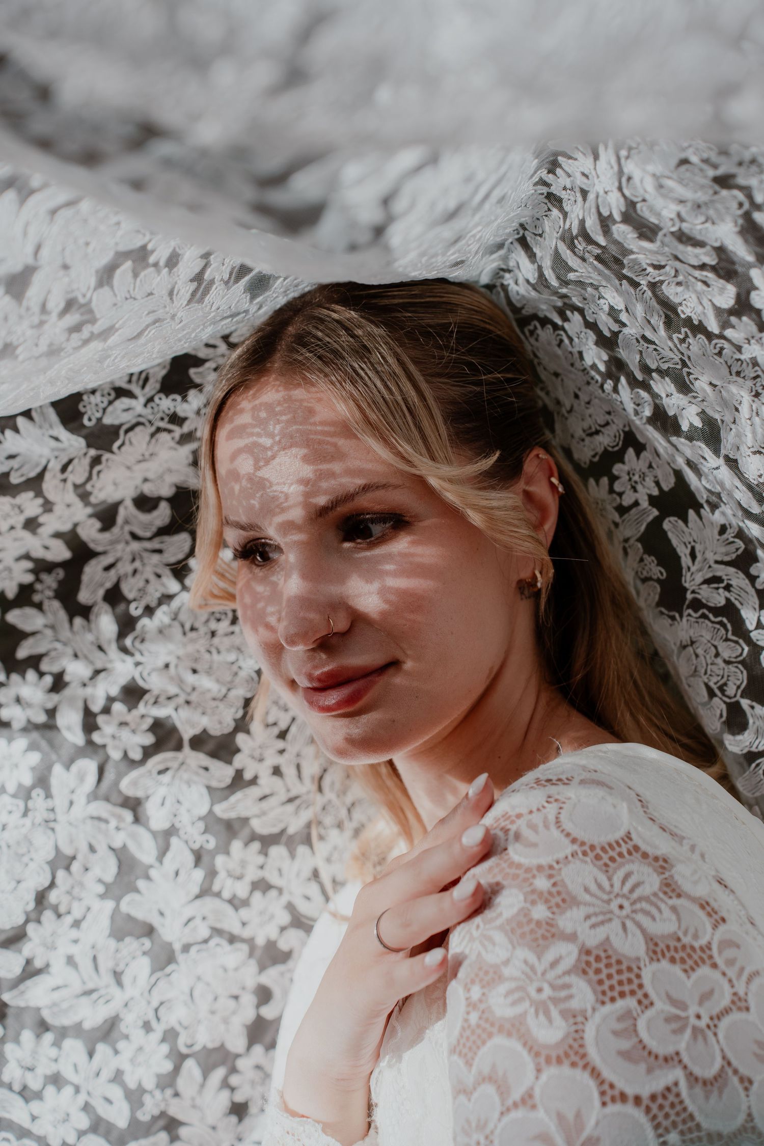 Bride in white lace dress poses under floral lace veil, with dappled light on her face, looking at camera.