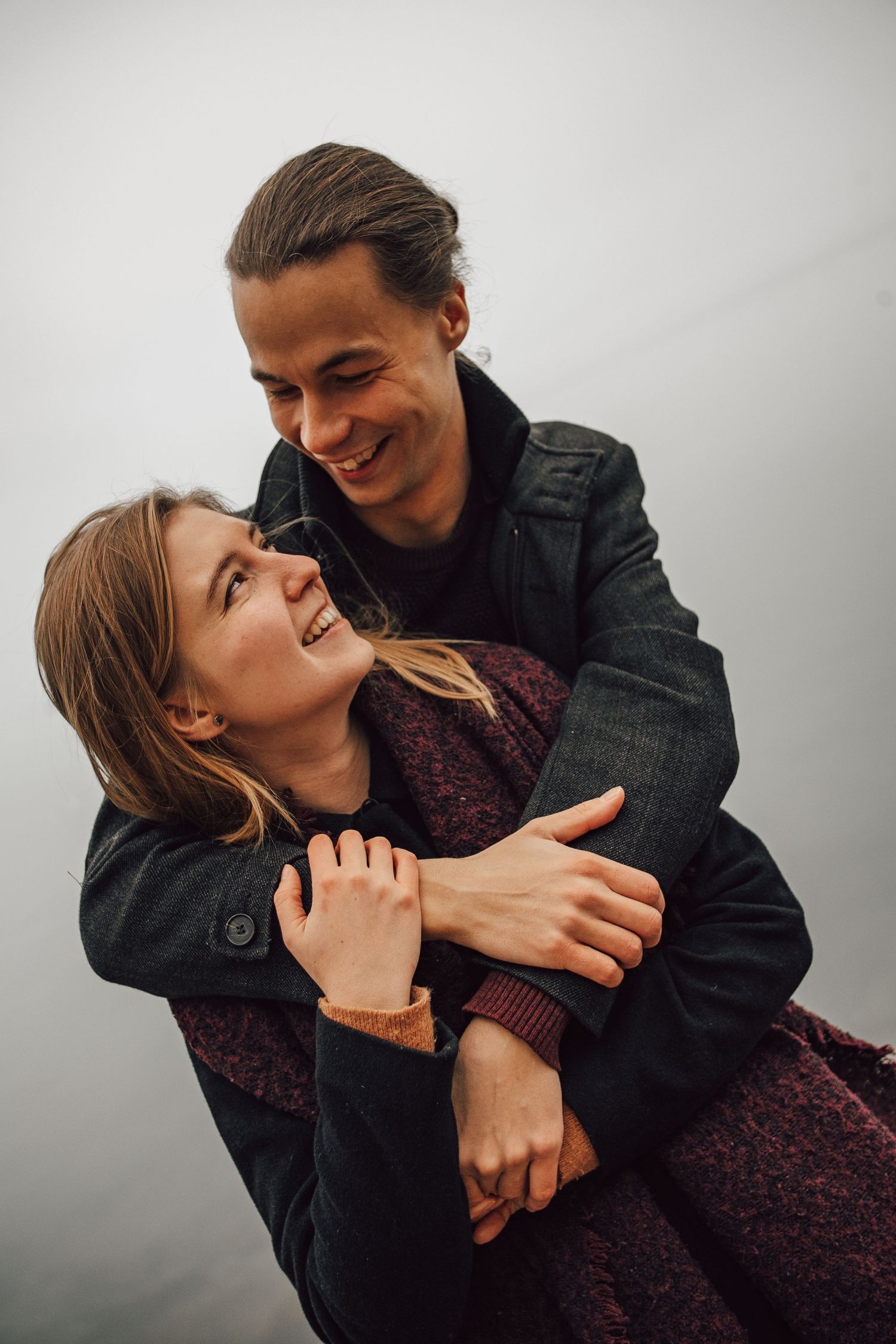 A romantic couple embraces and laughs together outdoors on a cloudy day, wearing dark winter coats.