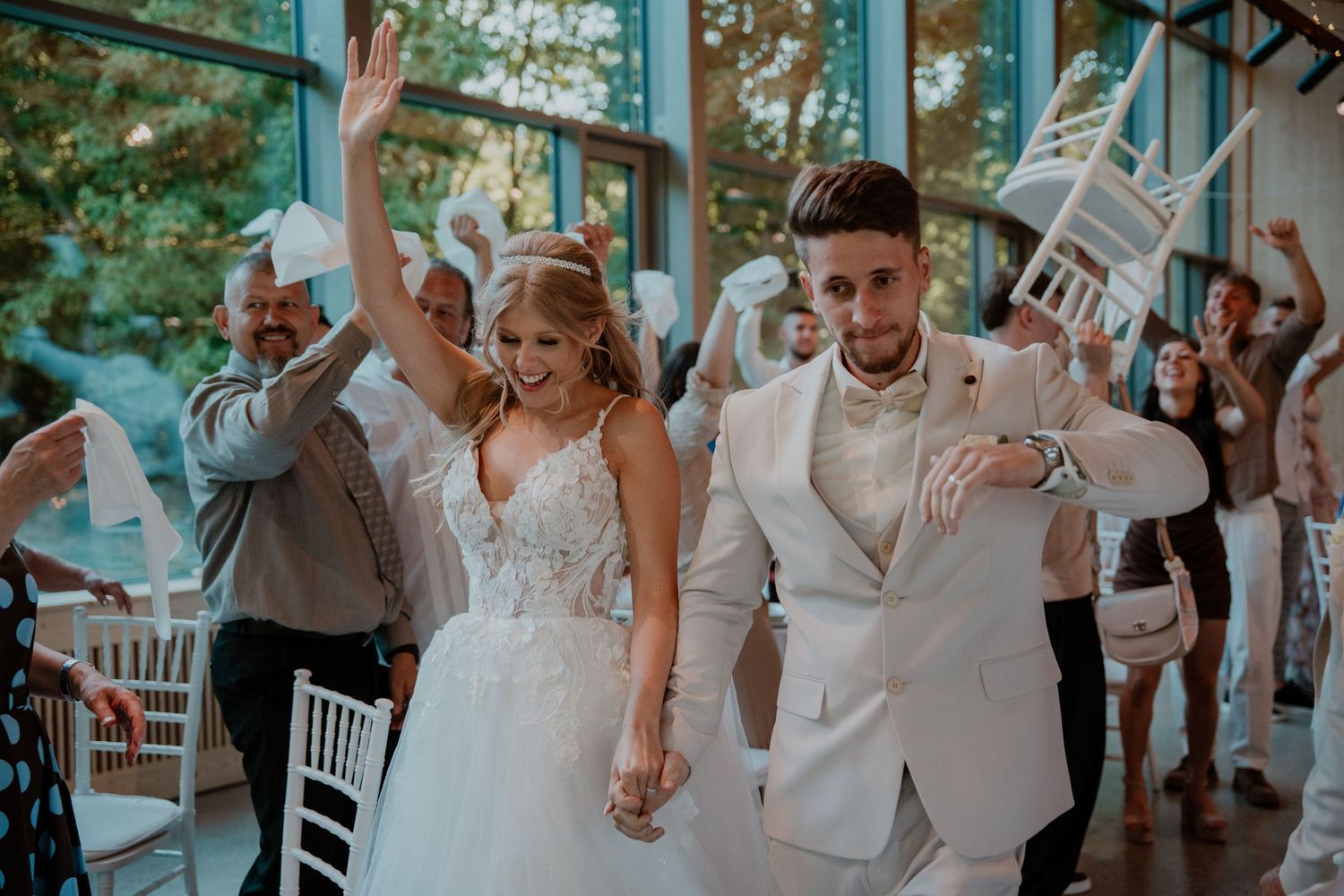 Bride and groom celebrate joyfully with guests raising chairs at elegant wedding reception venue.