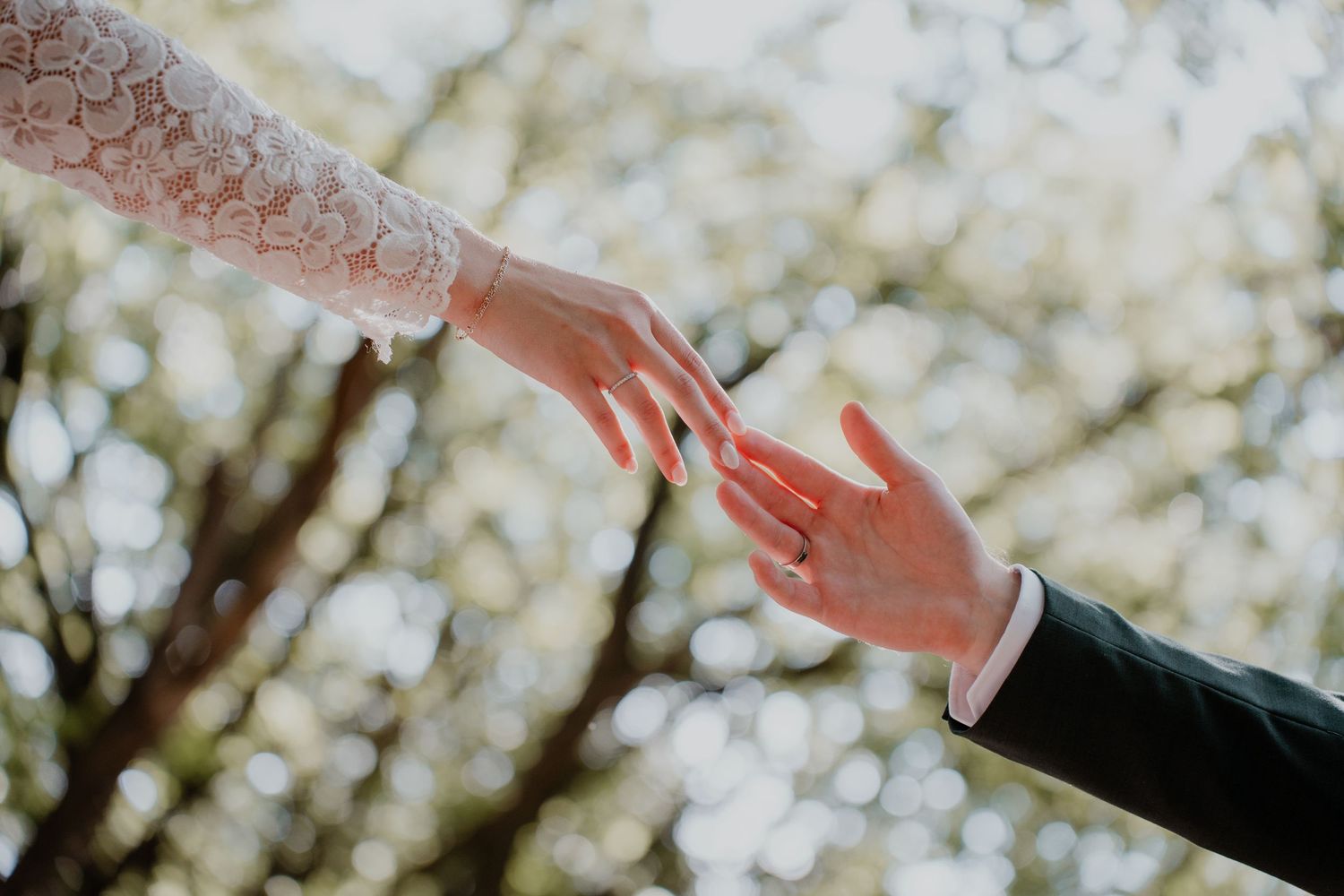 Bride and groom touching hands outdoors, with a wedding ring visible, surrounded by soft bokeh tree background.