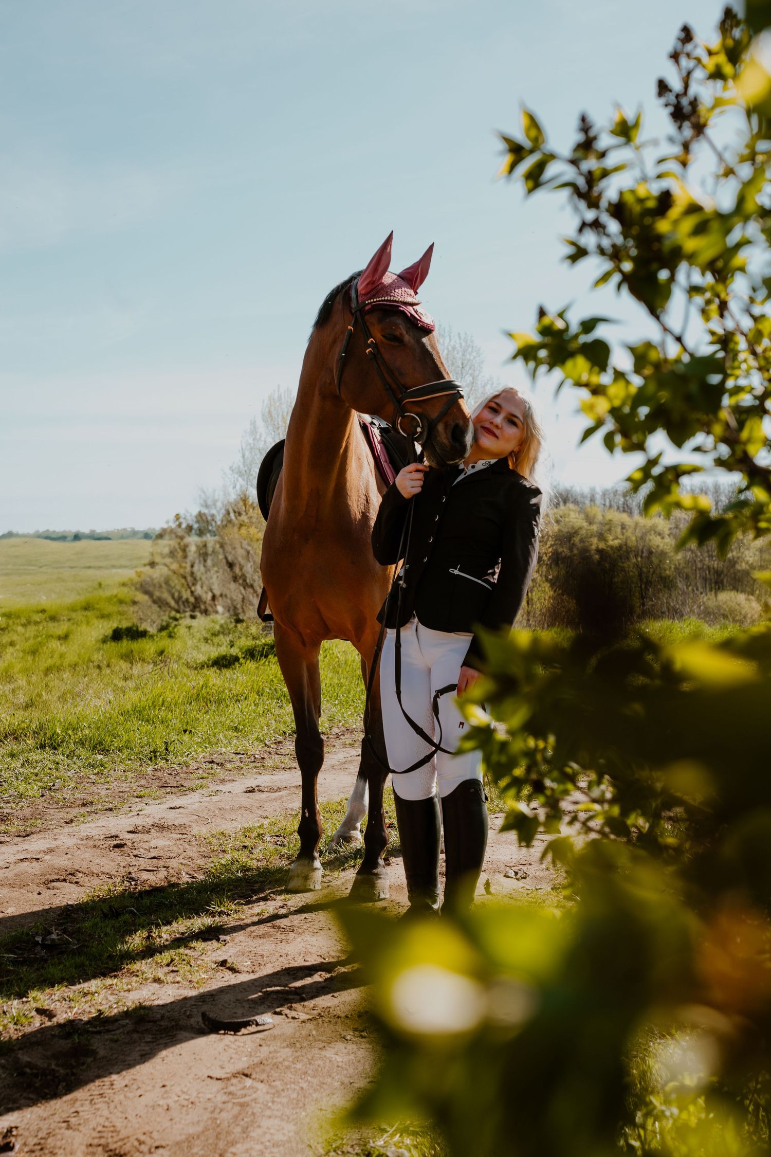 A beautiful chestnut horse stands with its rider on a dirt path surrounded by greenery on a sunny day.
