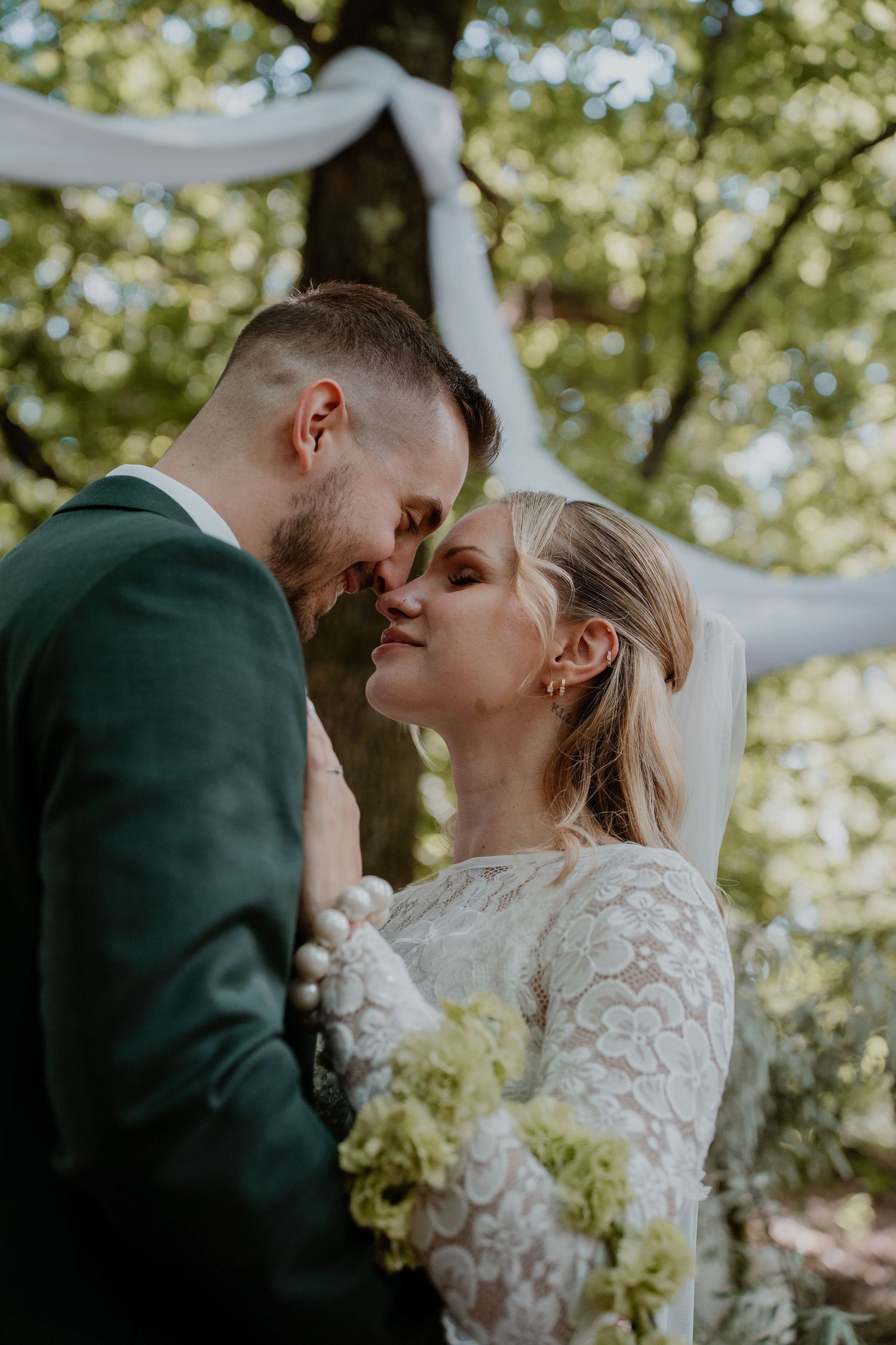A bride and groom share a romantic kiss outdoors under a tree during their wedding ceremony, she holds green hydrangeas.