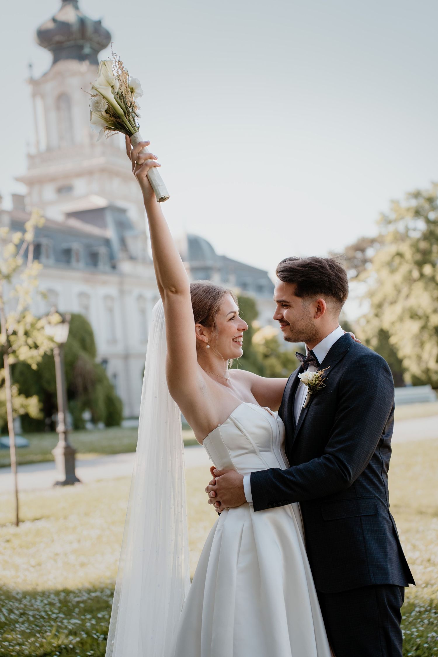 Joyful bride raises bouquet while groom embraces her in front of elegant building on their wedding day.