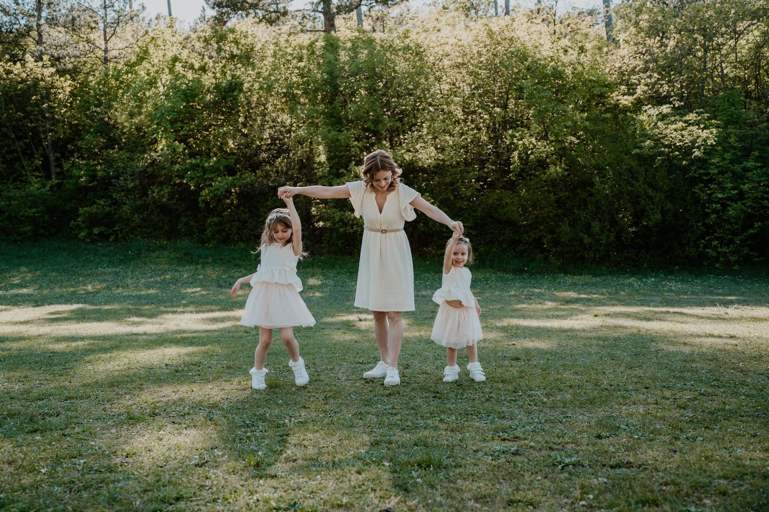 A mother in a white dress holds hands with two young girls in matching outfits, dancing together on a green lawn.