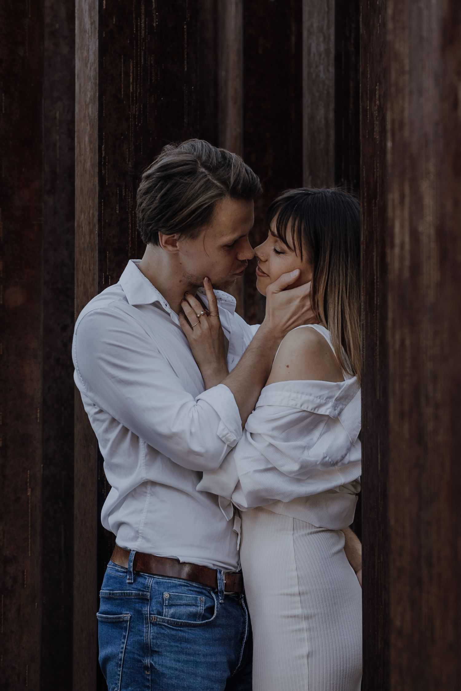 A romantic couple in blue jeans and white tops embrace against a dark wooden wall background.