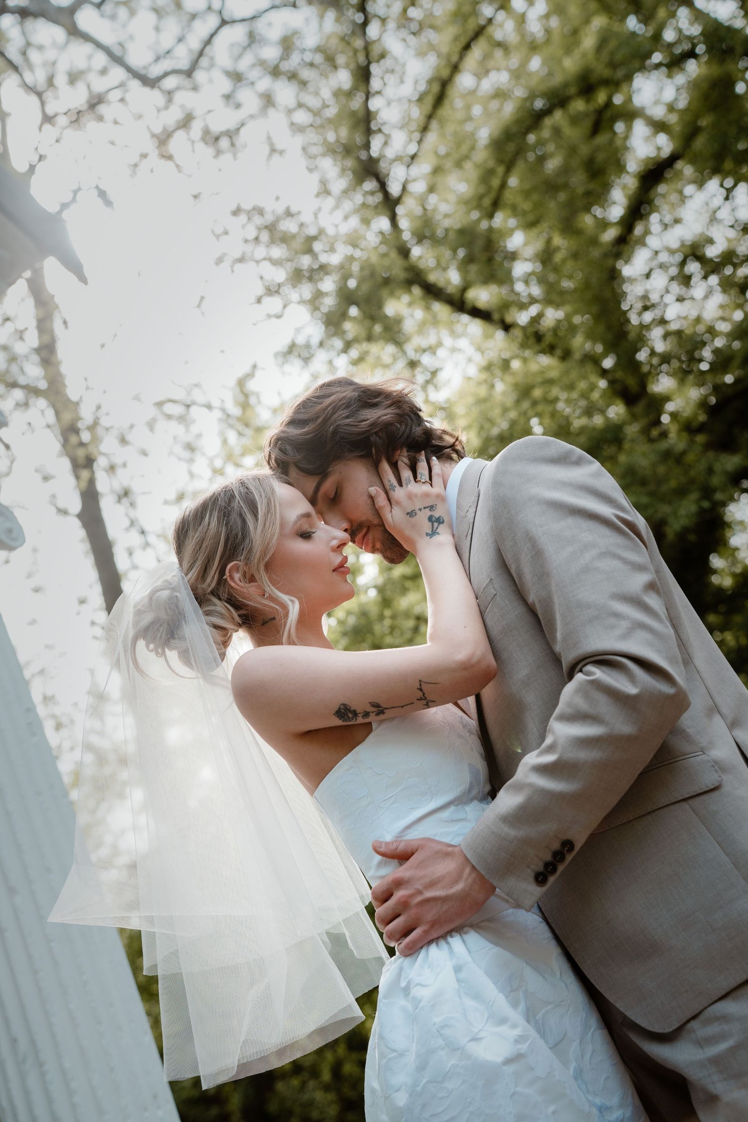 Bride and groom share a romantic almost-kiss outdoors, bride in white dress with veil, groom in tan suit, surrounded by trees.