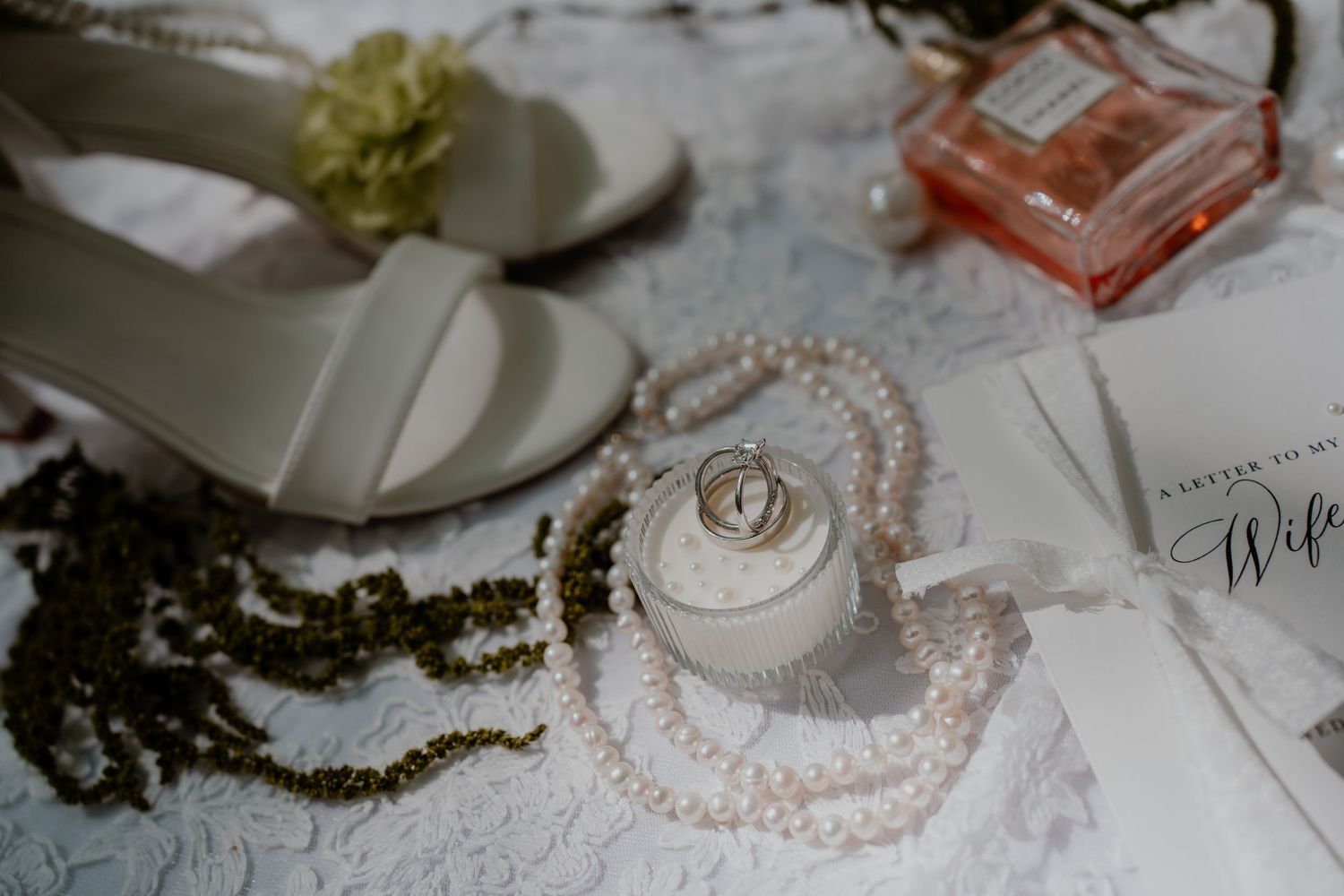 Wedding flat lay with white heels, pearl necklace, gold rings, perfume bottle, and a Wife card on lace fabric.