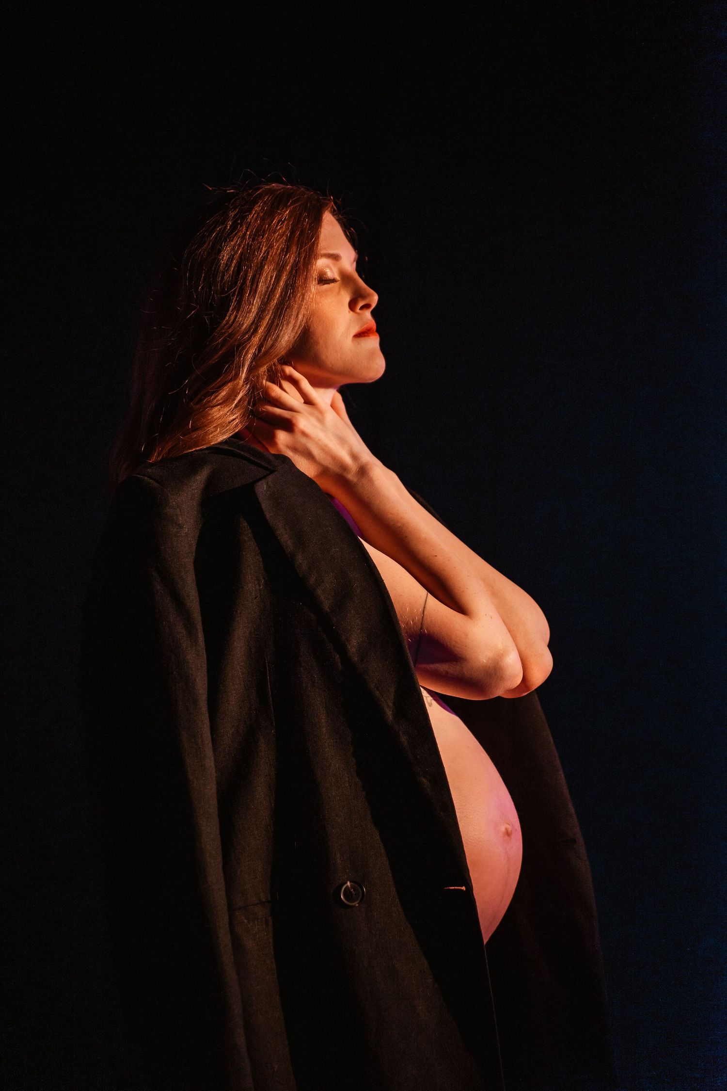 A side profile silhouette shows a pregnant belly beneath a dark coat against a black background with dramatic lighting.