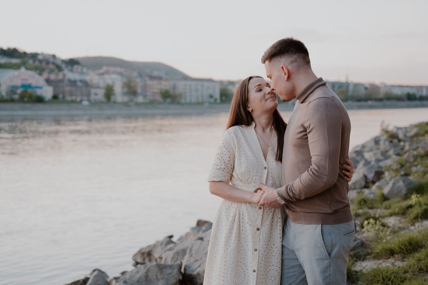 Romantic couple sharing a tender moment by a riverside at sunset, woman in white dress embraced by man in beige sweater.