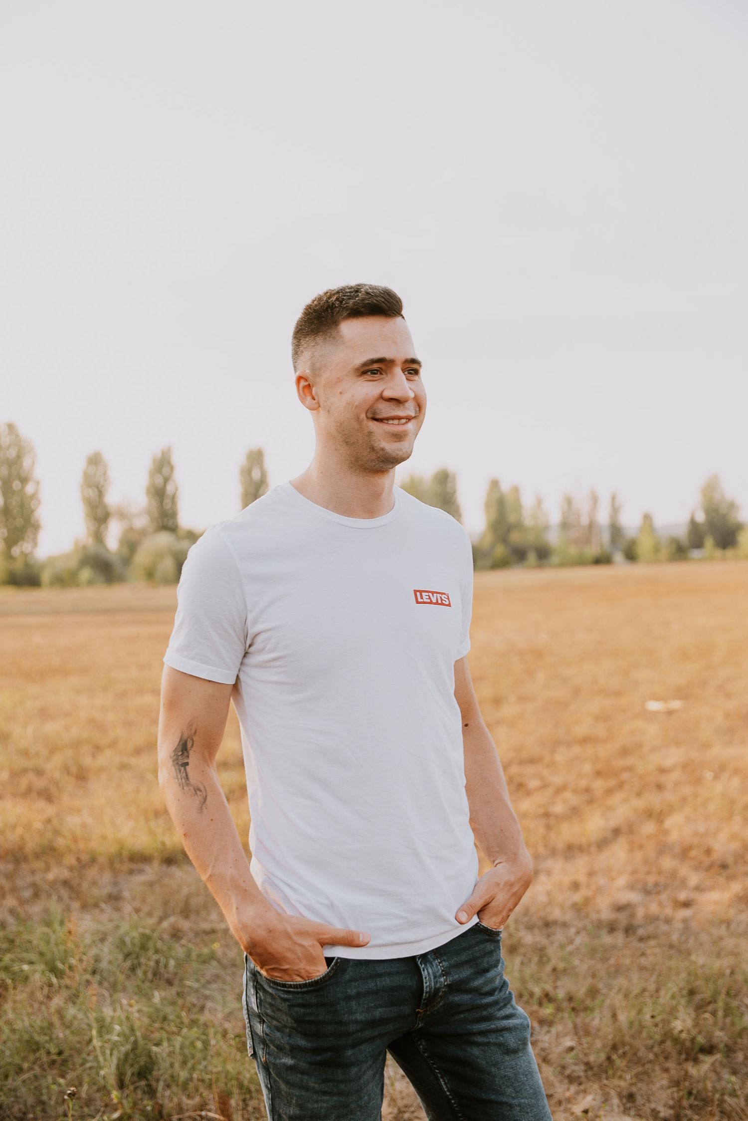 Person in a plain white t-shirt and jeans stands smiling in a golden field at sunset with trees in the distance.