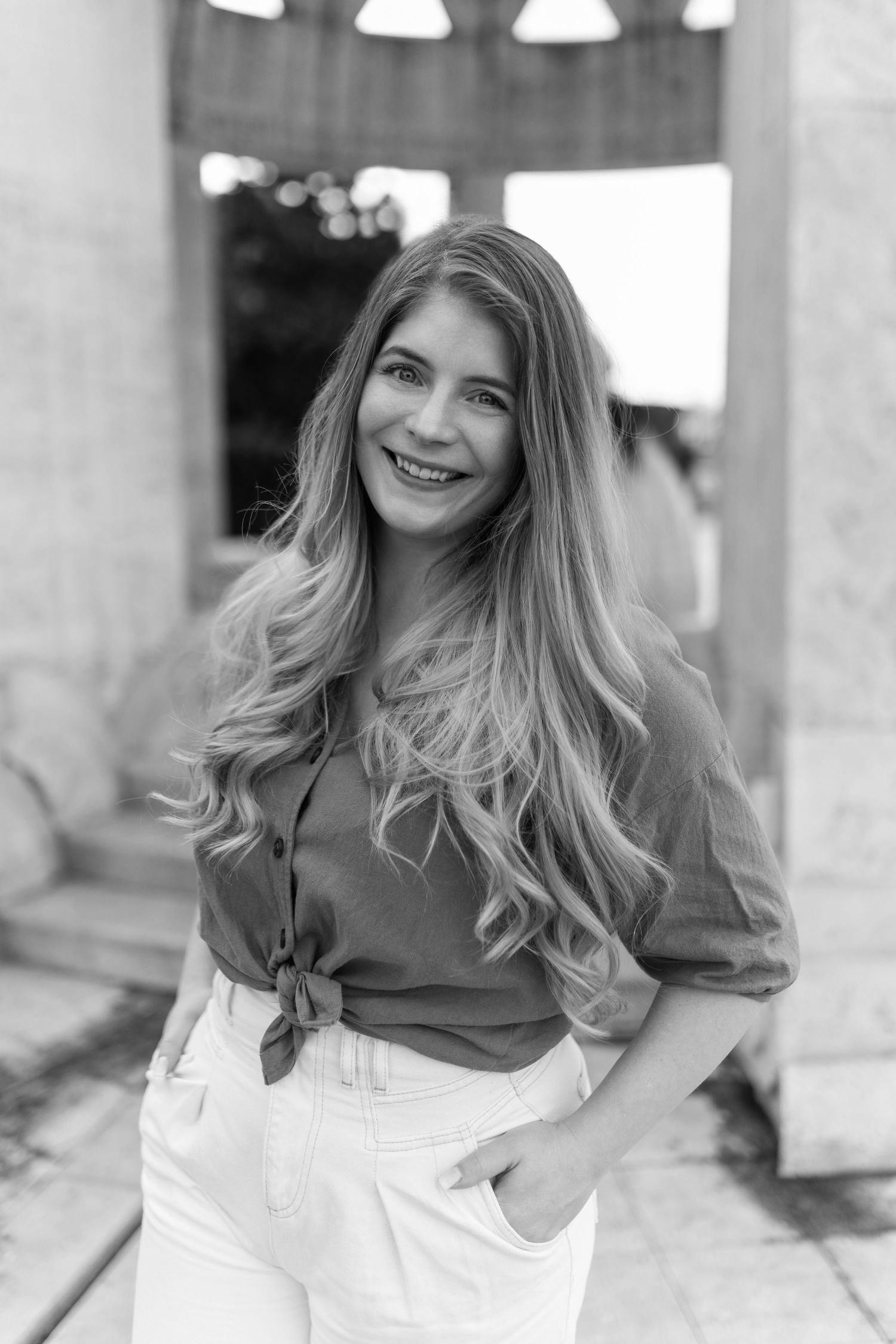 Black and white portrait of a smiling young woman with long wavy hair, wearing a tied blouse and white pants outdoors.