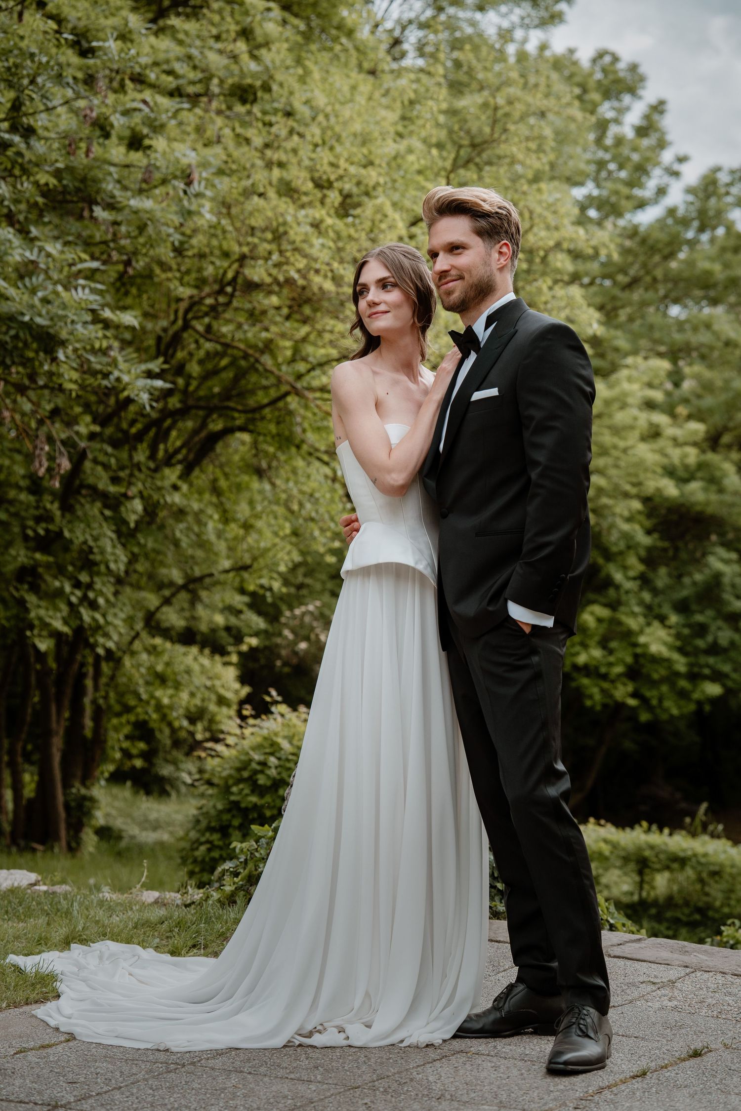 Elegant bride in white strapless gown and groom in black tuxedo posing together in a lush green garden.