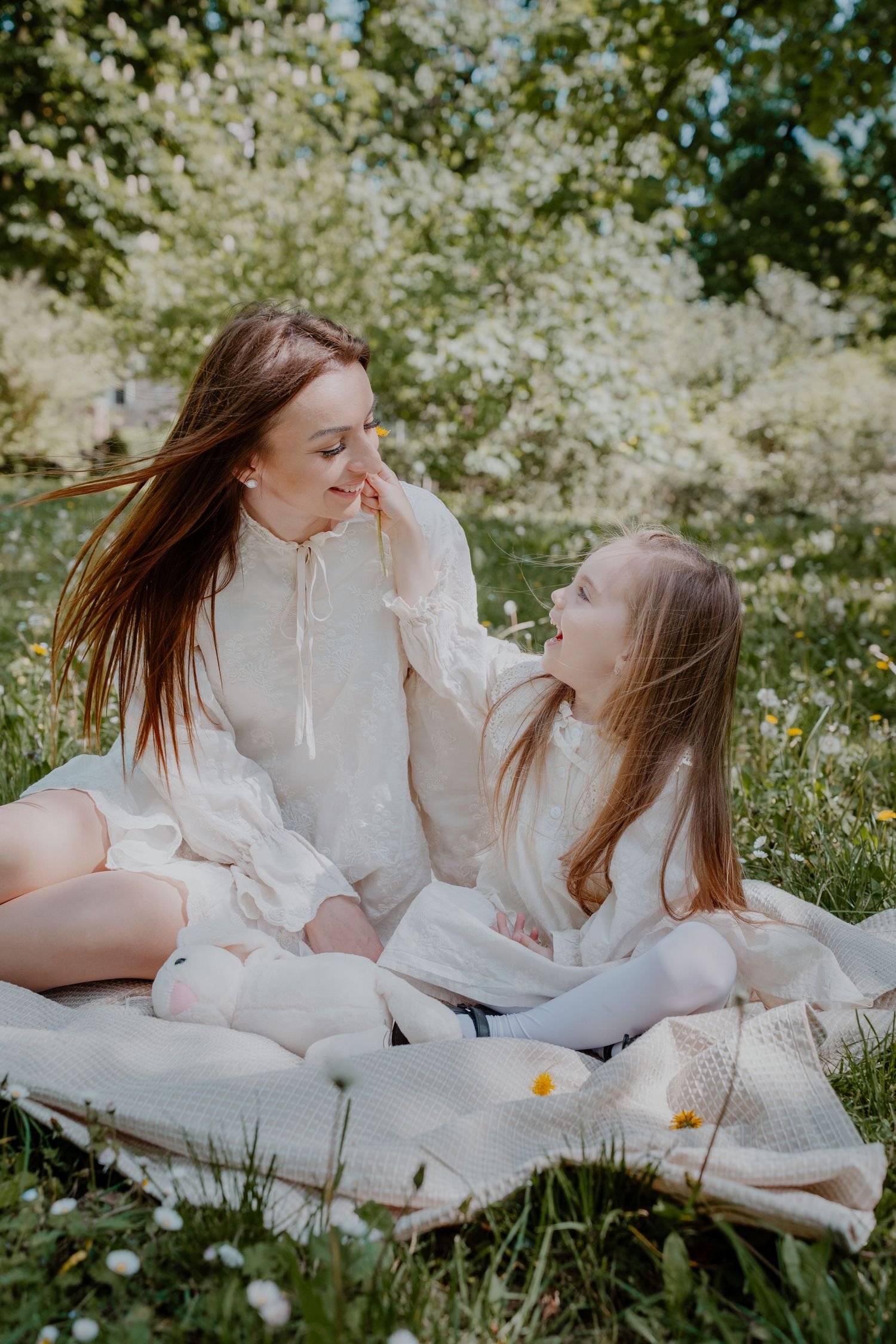 A mother and daughter in matching white dresses share a tender moment on a blanket in a blooming spring garden.