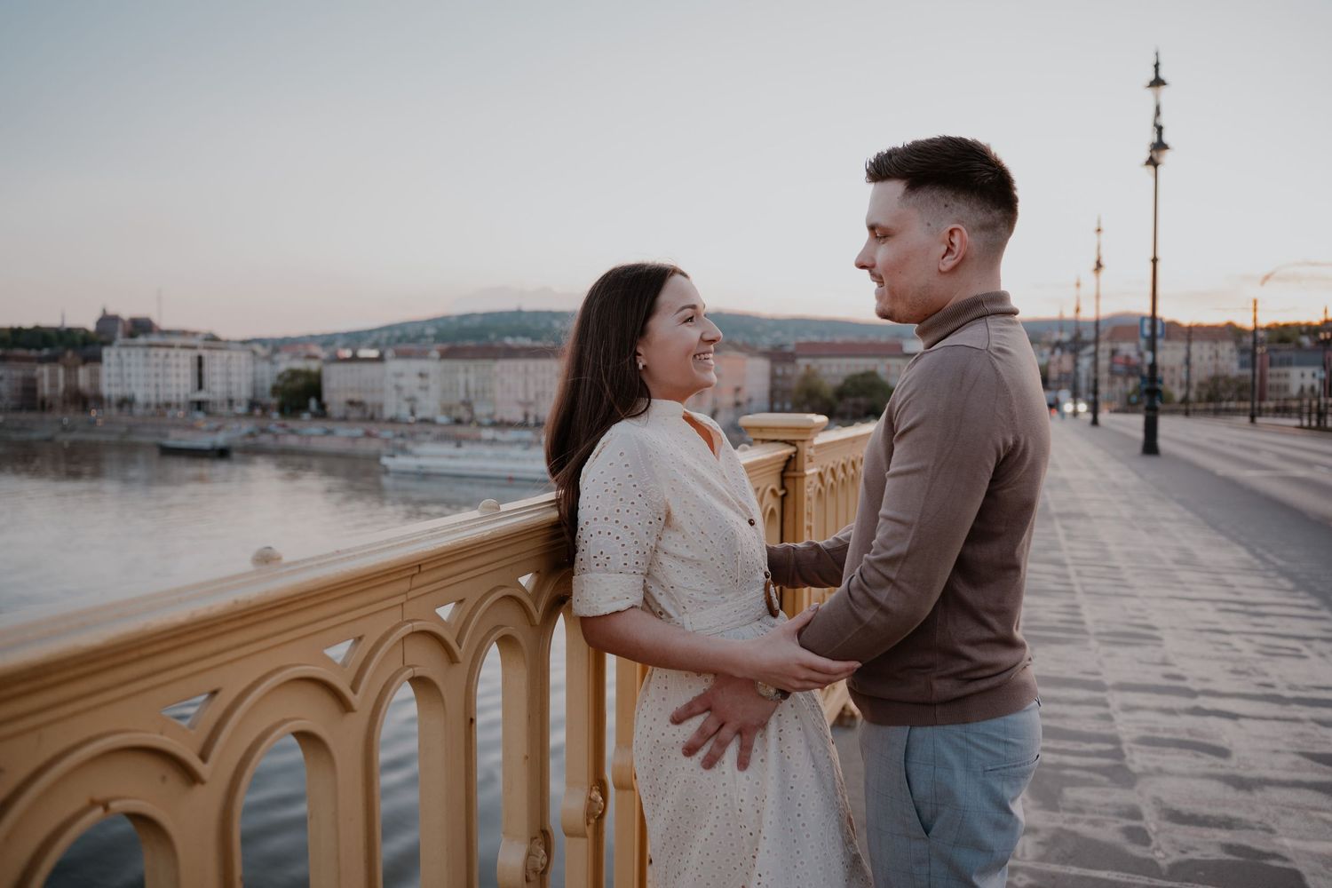 Couple holding hands on a scenic bridge at sunset with a river and city skyline in the background.