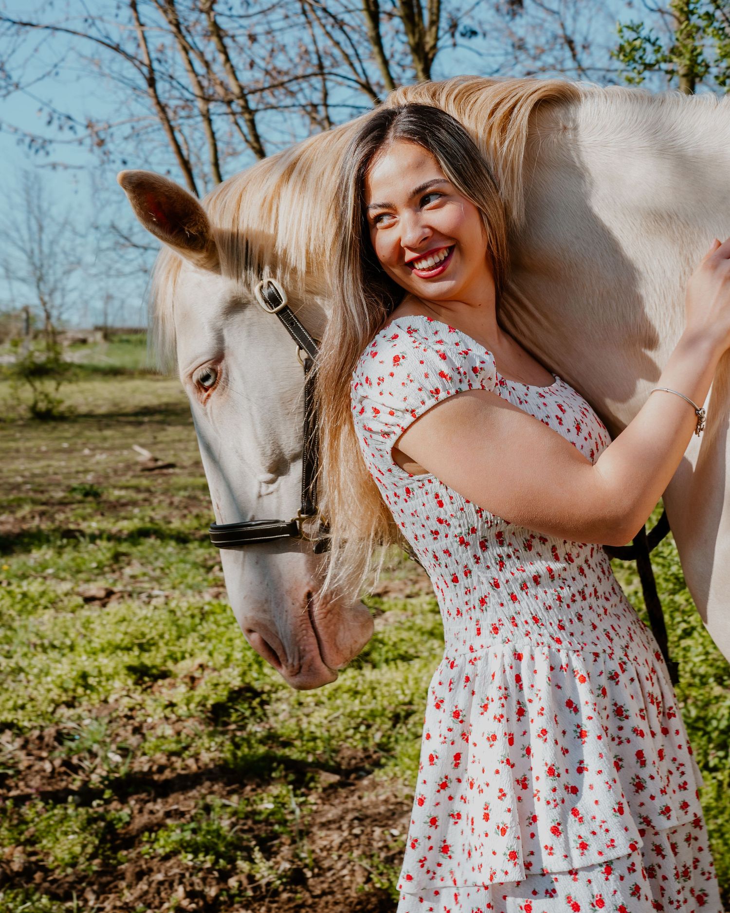 Smiling person in a floral dress stands next to a white horse in a sunny outdoor setting with trees in the background.