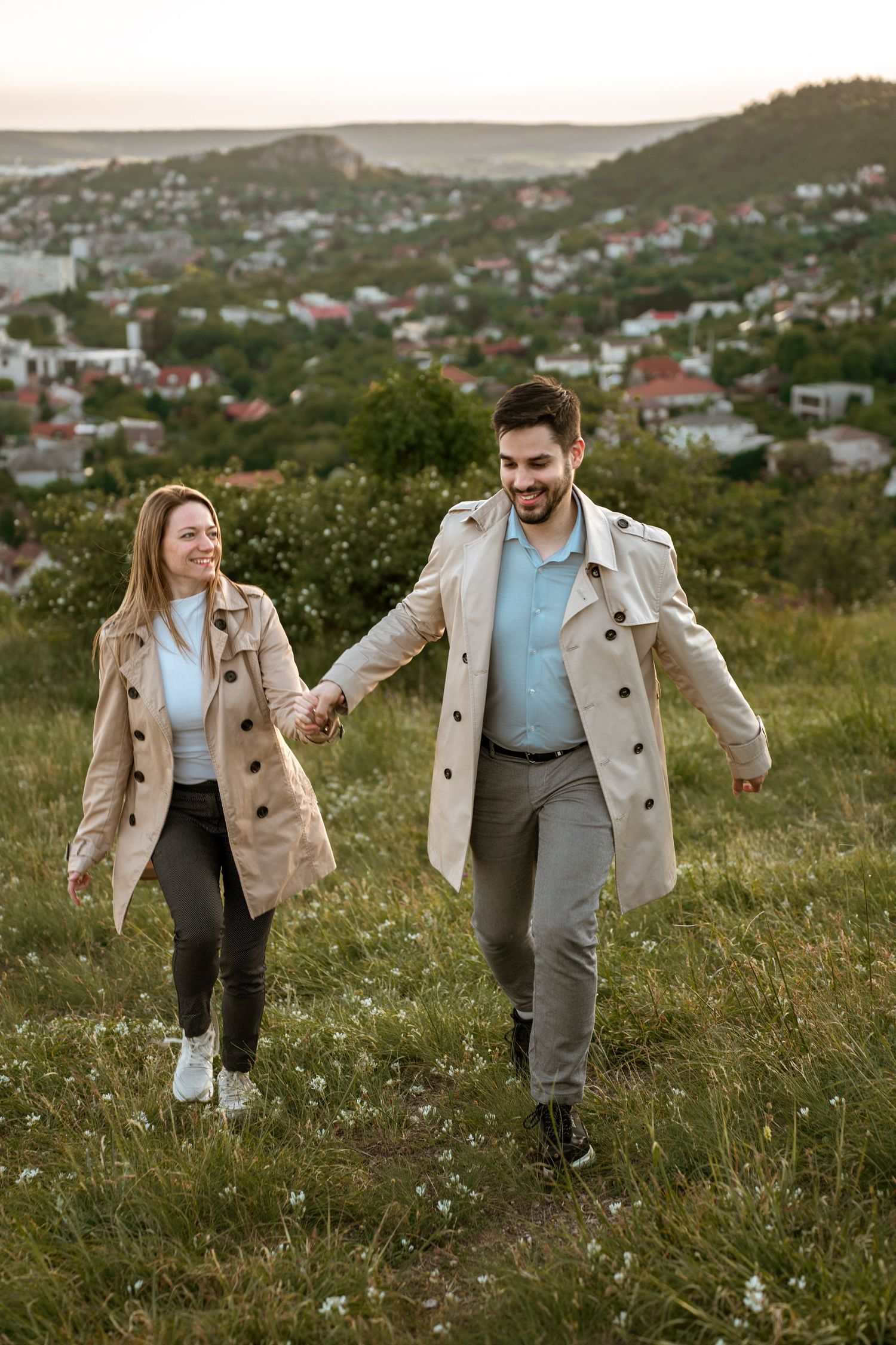 A couple in matching beige trench coats walk hand in hand on a grassy hillside overlooking a suburban town at dusk.