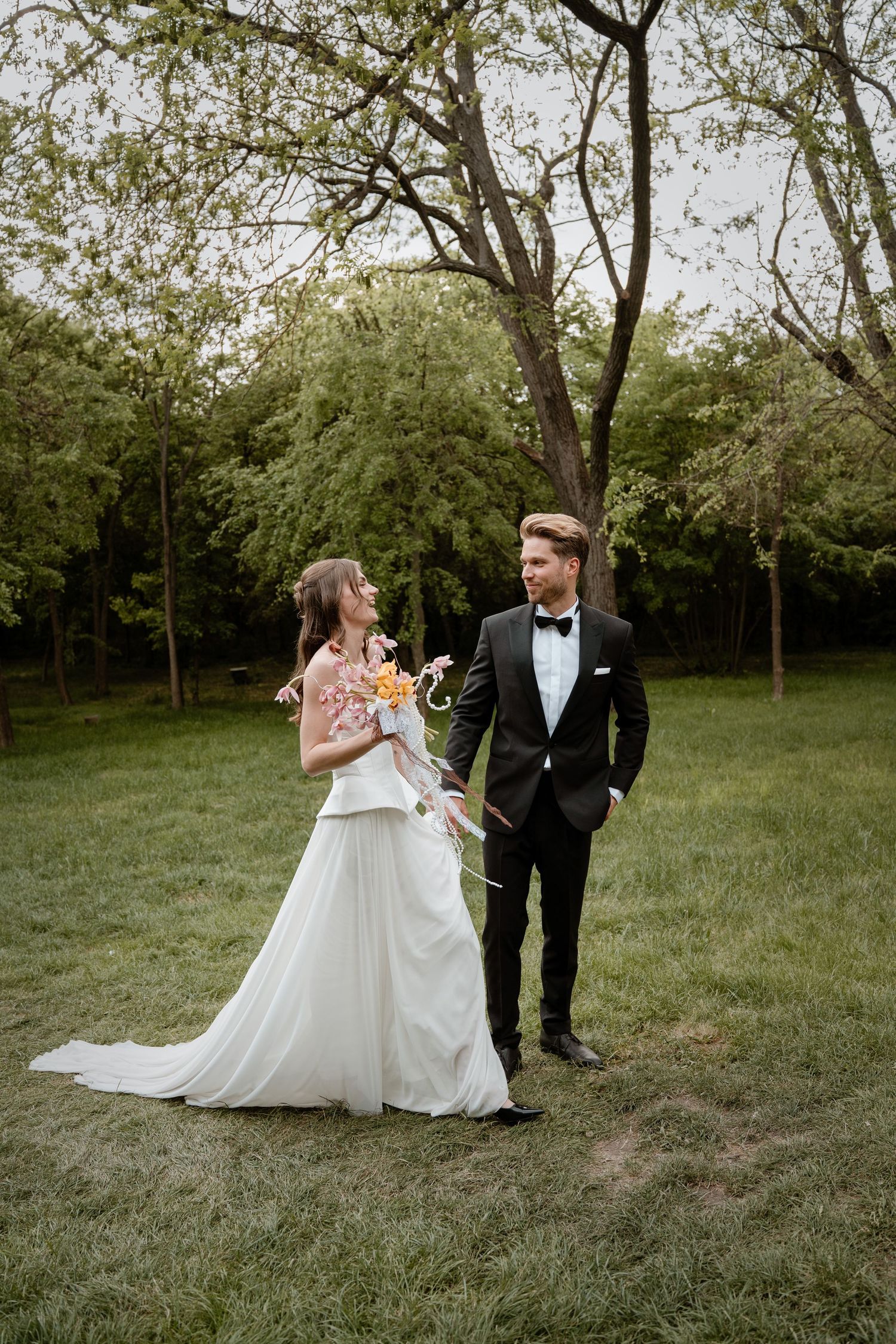 A bride in a white gown holding a bouquet stands with a groom in a black tuxedo in a lush green outdoor setting.