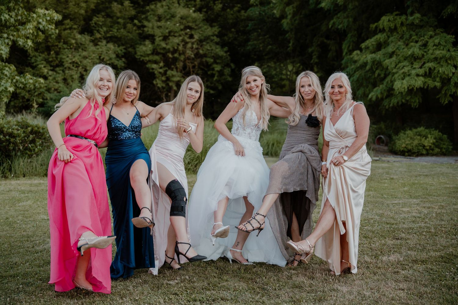 Bride poses with six bridesmaids in colorful gowns, laughing and kicking up heels on a green lawn.
