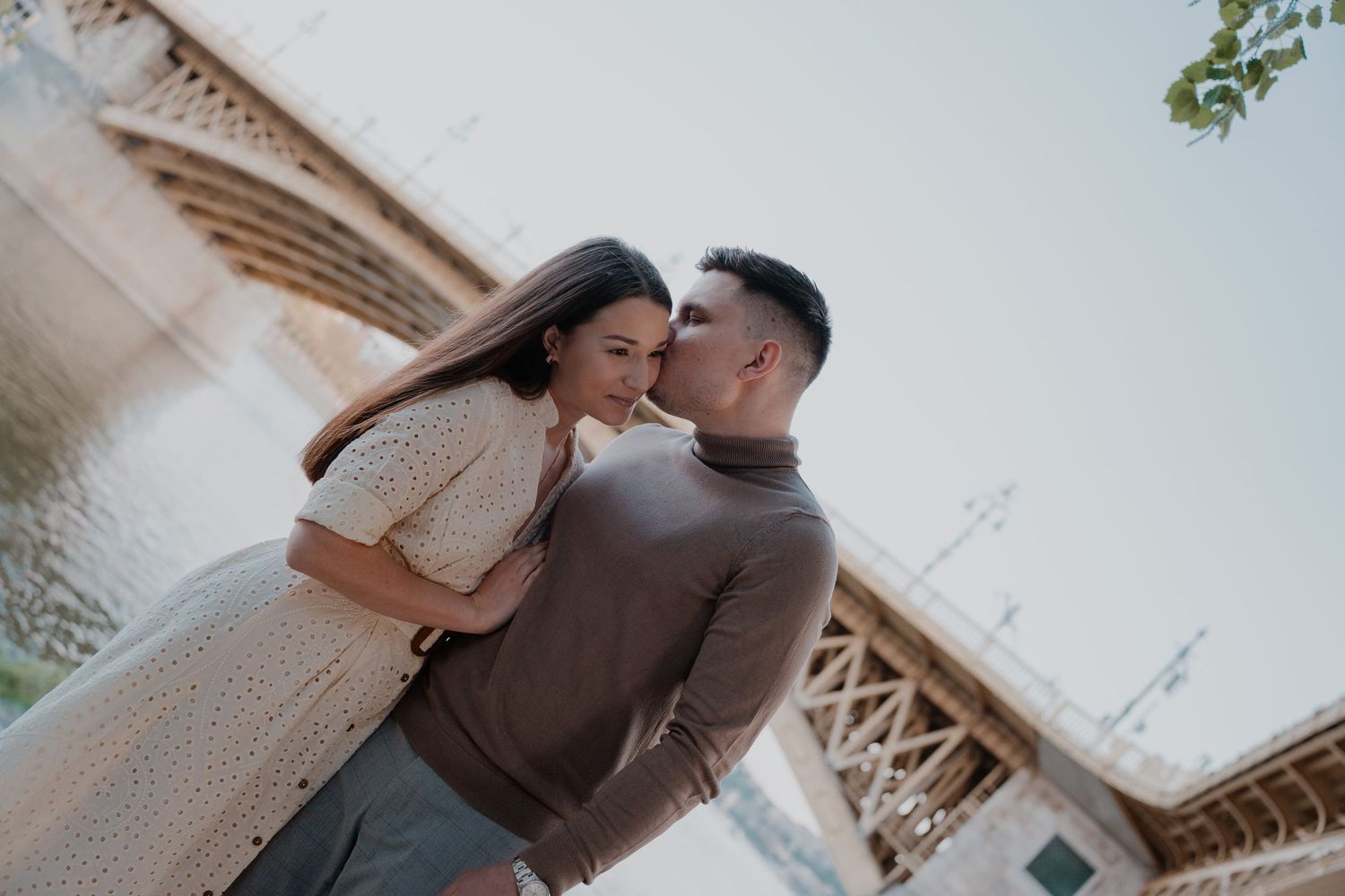 Romantic couple sharing a tender moment near a river bridge on a sunny day, perfect for love and relationship themes.