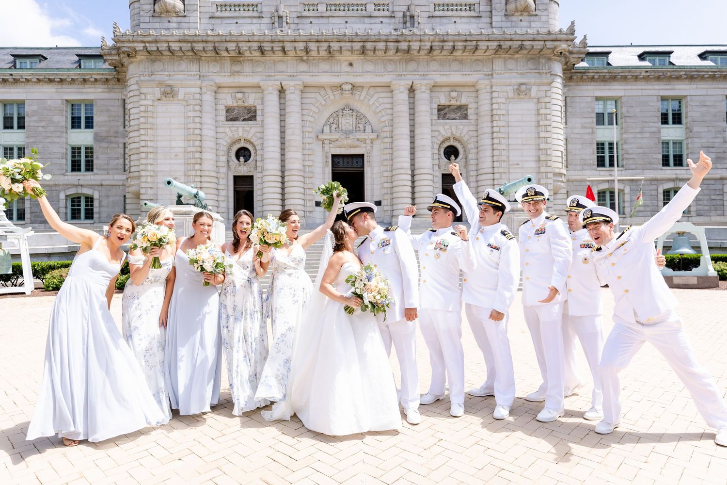 Wedding party in white dresses and uniforms poses joyfully in front of ornate stone building architecture.