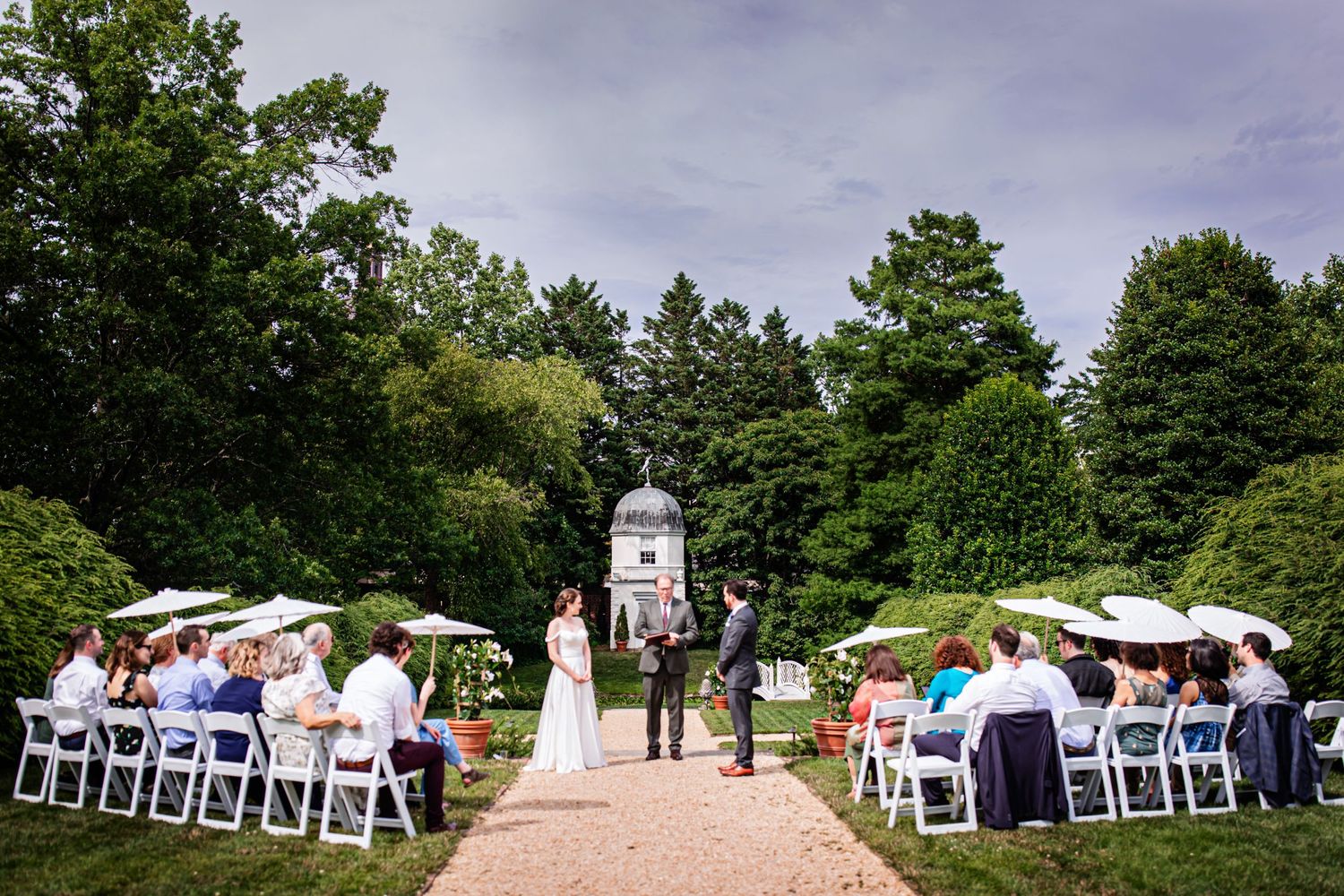 Wedding ceremony site with white chairs lined up along garden path leading to white gazebo.