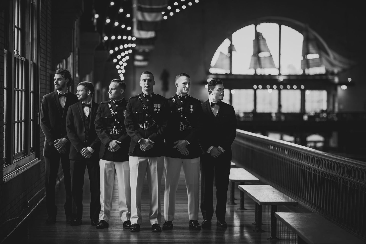 Groomsmen in formal attire pose by arched windows with string lights.