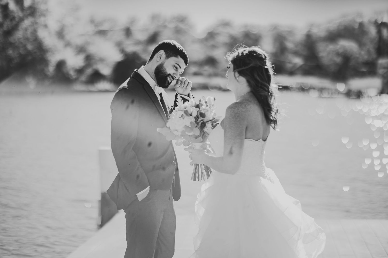 A black and white waterfront wedding photo with a couple sharing a joyful moment.