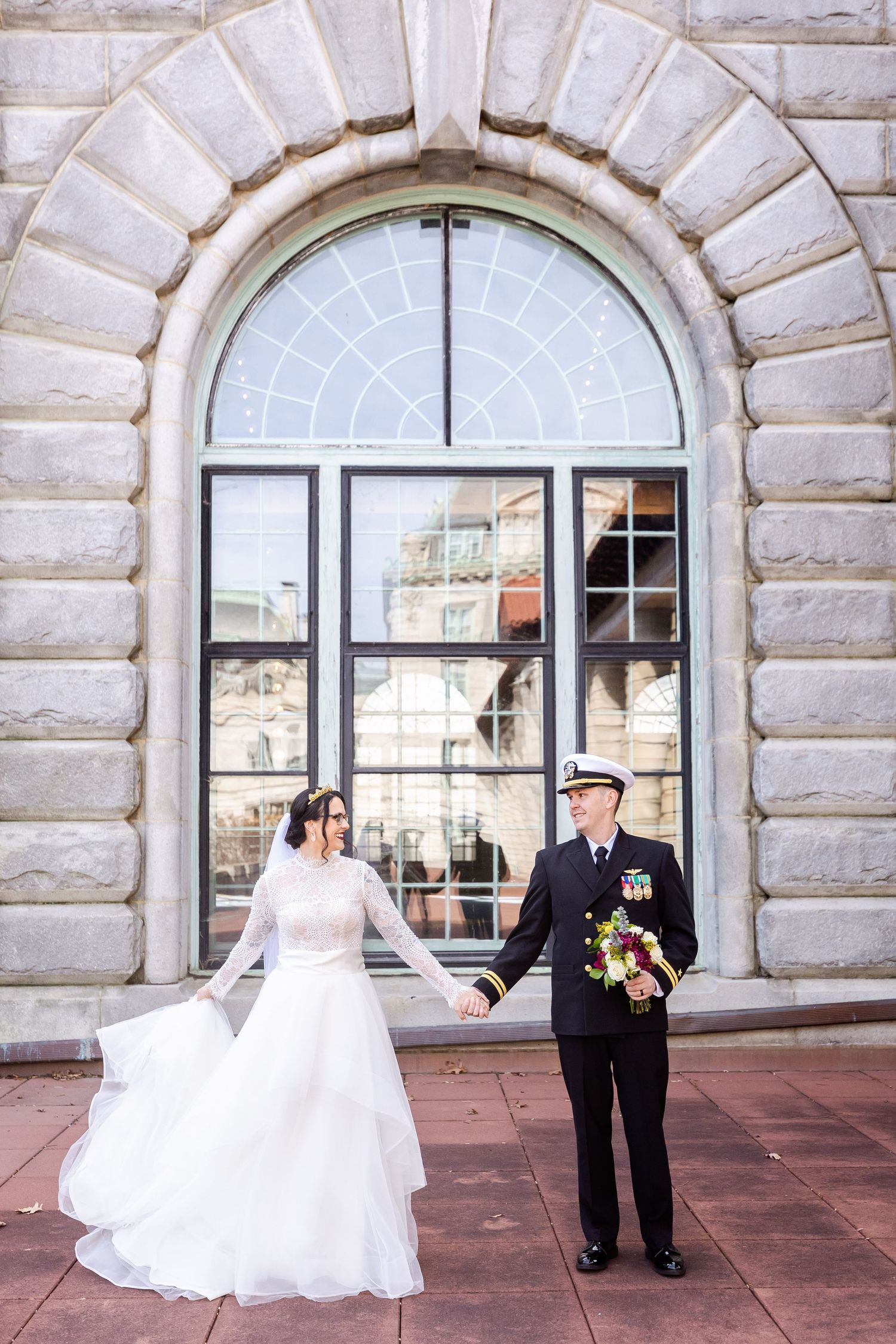 Wedding couple posing in front of an elegant arched window at a historic stone building.