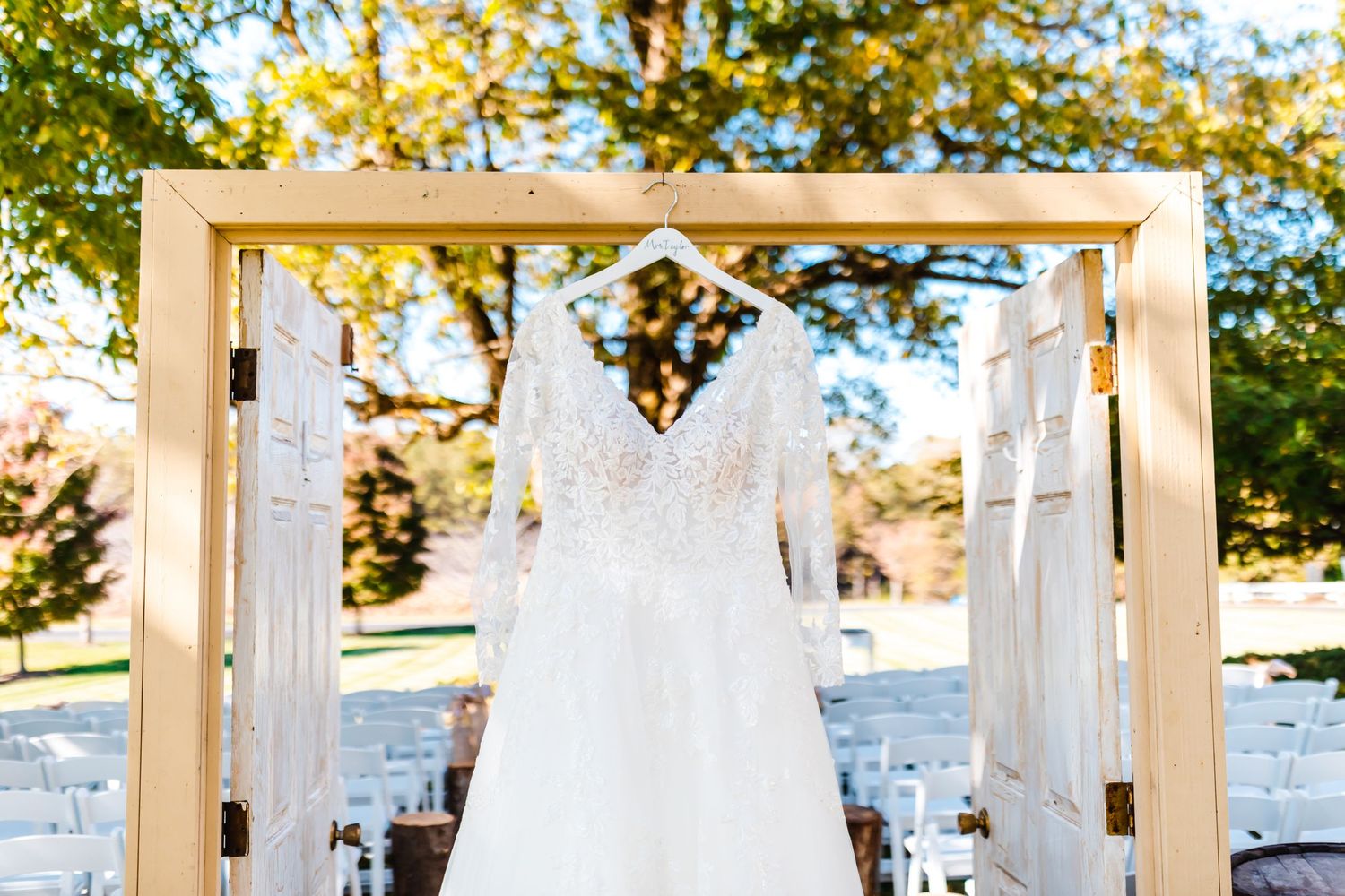 White lace wedding dress displayed on wooden arch with reception chairs in background.