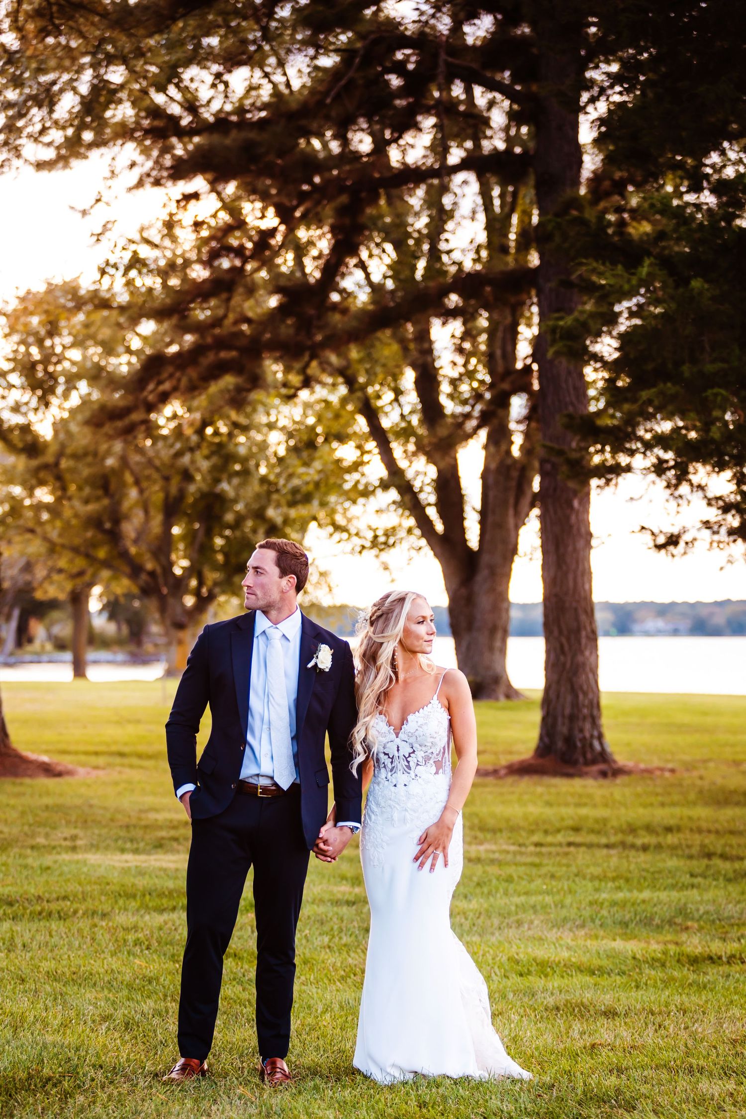 Sunset wedding portrait with couple walking near waterfront and tall trees.