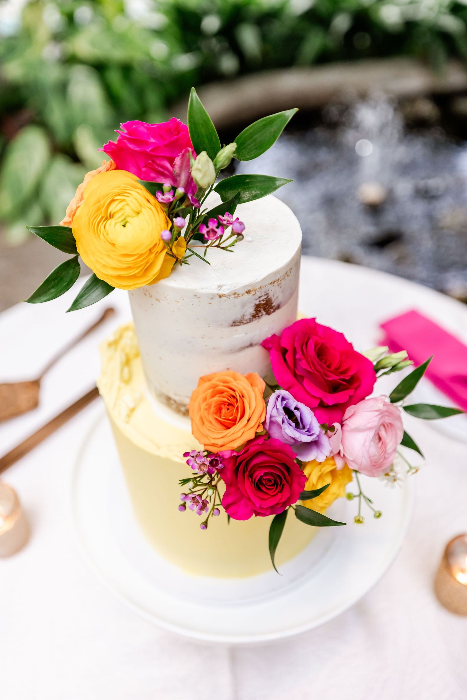 Elegant two-tier wedding cake decorated with vibrant yellow, pink, and purple fresh flowers on a white plate.
