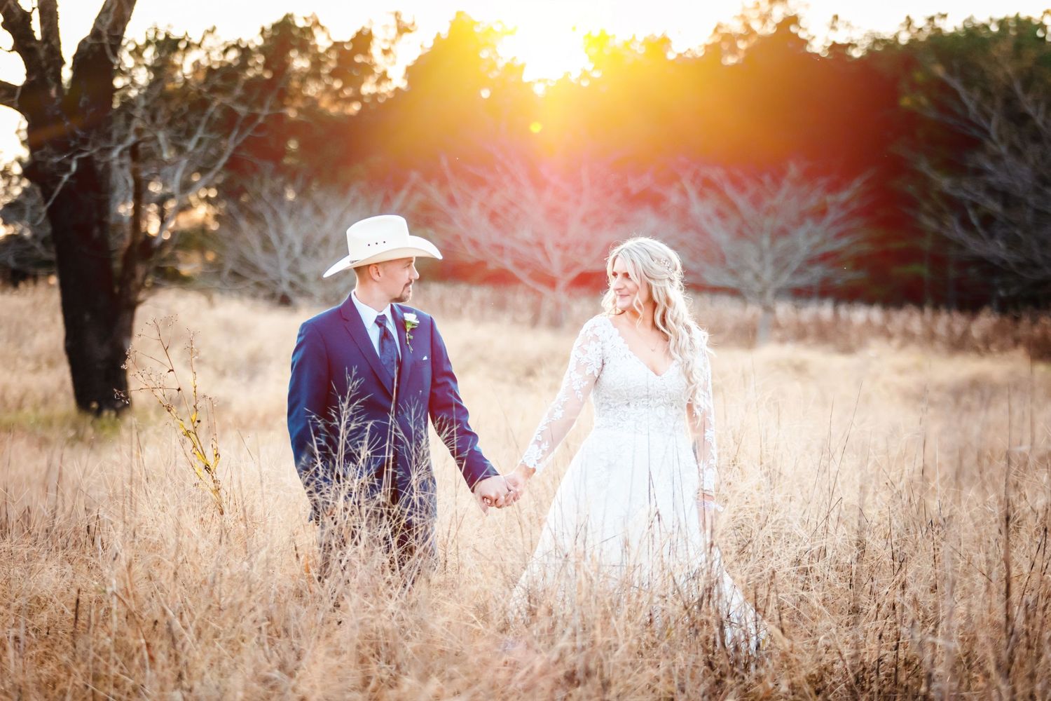 Couple walks hand in hand through golden field at sunset wearing wedding attire.