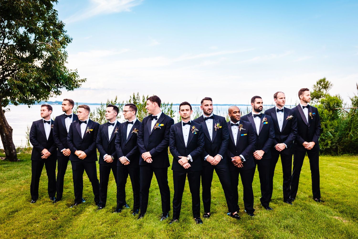 Groomsmen in black tuxedos and bow ties pose together on green lawn.