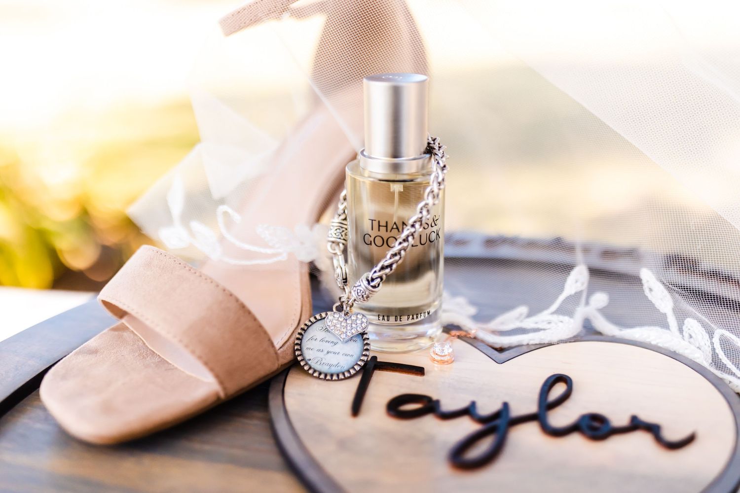 Wedding accessories including blush shoes, perfume bottle and personalized name tag on grey surface.