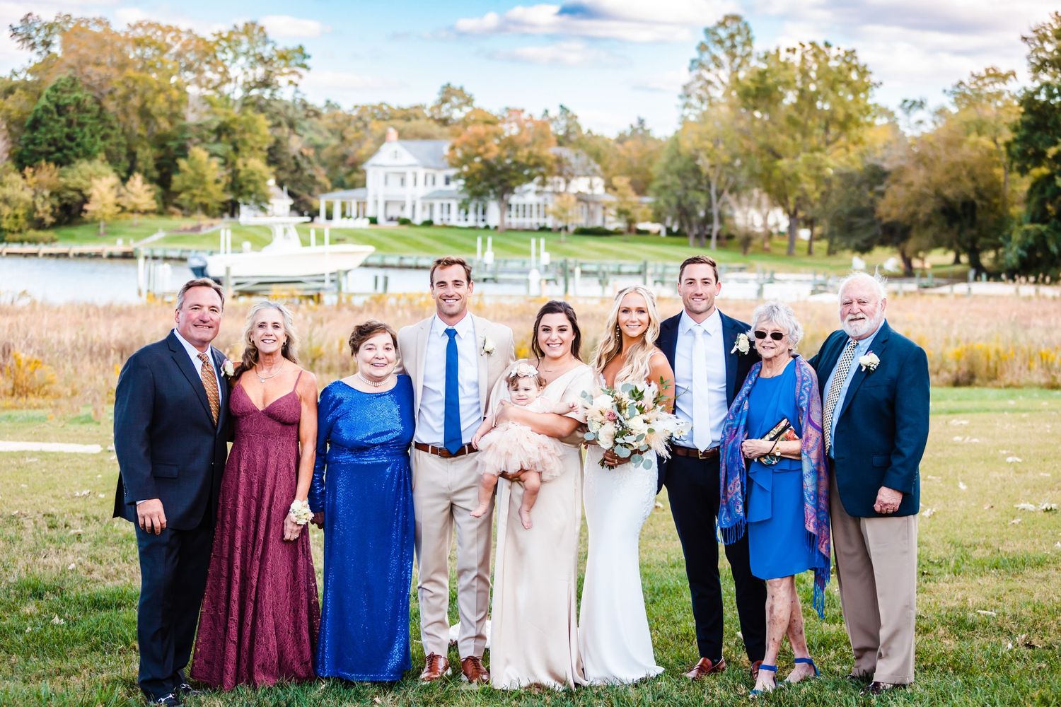 Wedding party poses for formal photo in front of rustic farmhouse and autumn field.