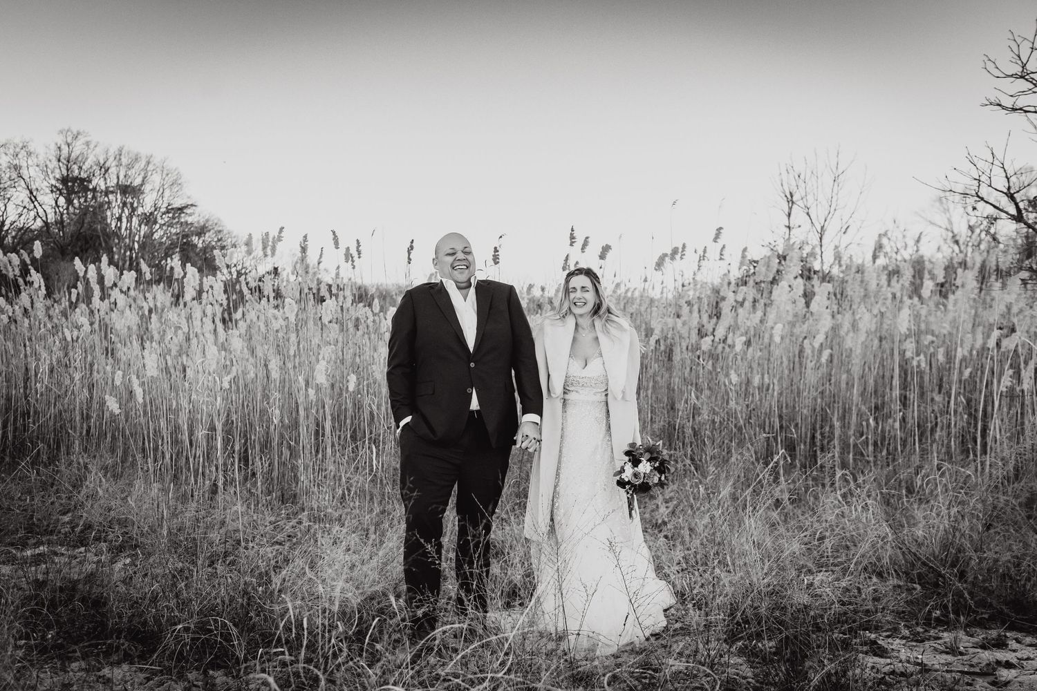 Black and white portrait of newlyweds standing in tall grass during golden hour.
