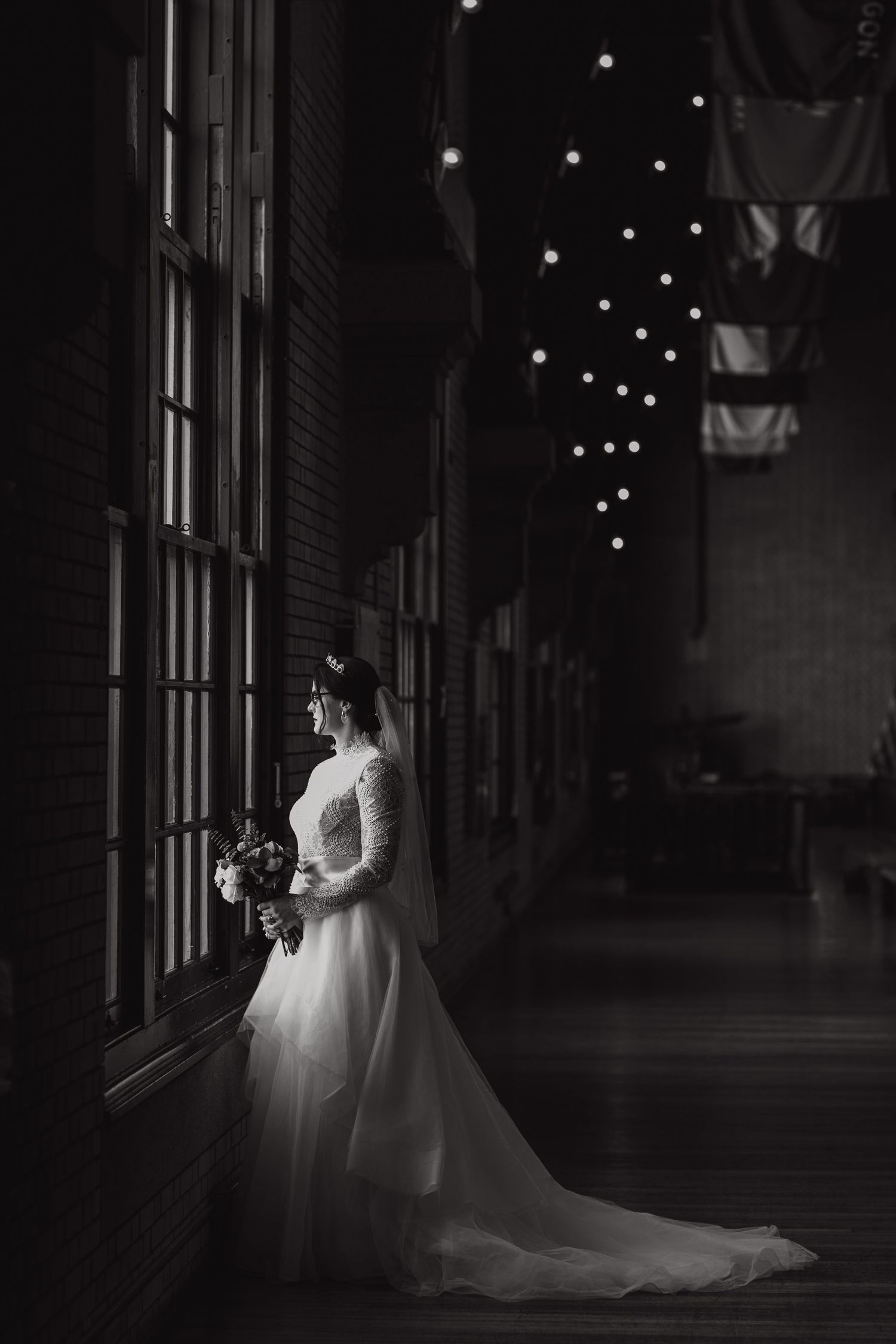 Dramatic black and white portrait of a bride in lace gown by window with string lights.
