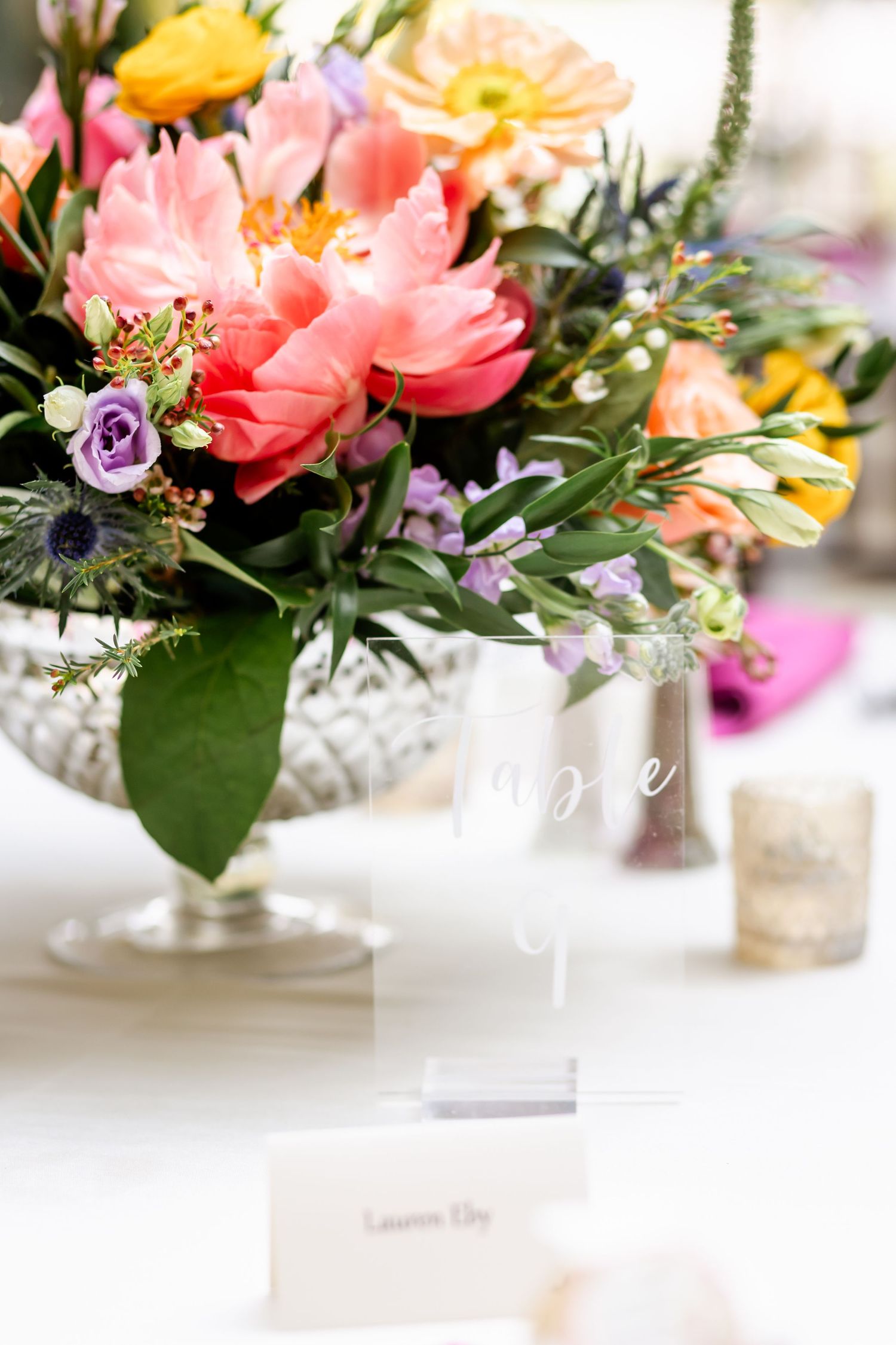 Colorful floral centerpiece with coral peonies, purple blooms and greenery in a silver pedestal vase.