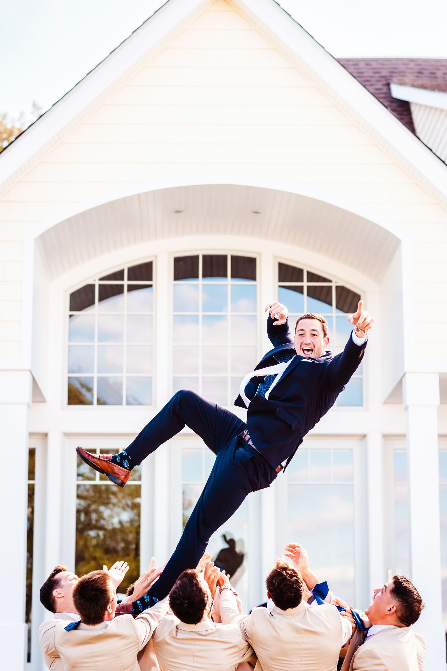Groomsmen toss groom in the air in front of a white chapel with arched windows.