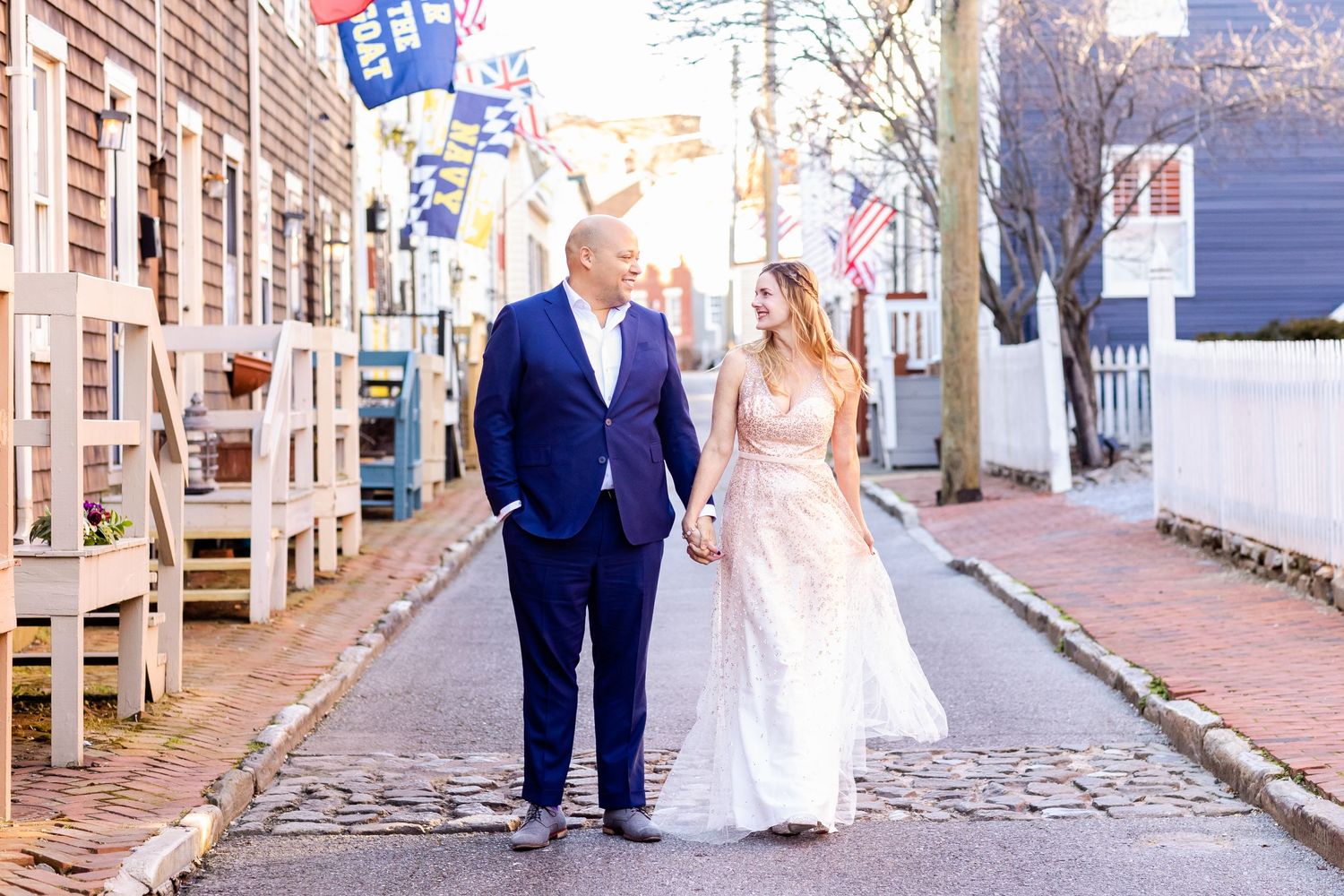 Couple walking down historic cobblestone street with colonial buildings and flags.