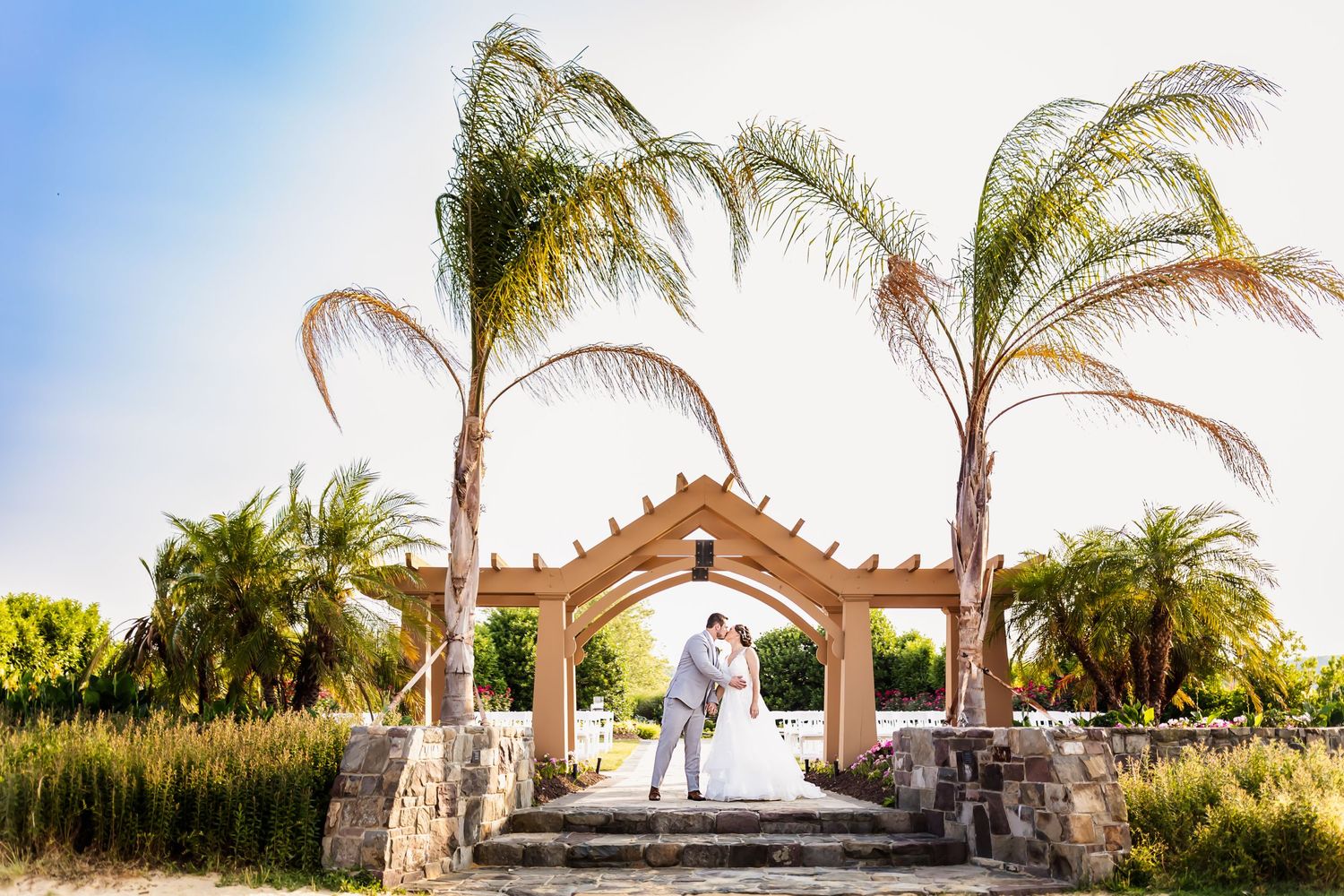 Wedding couple under a stone archway with palm trees and blue sky in a tropical setting.