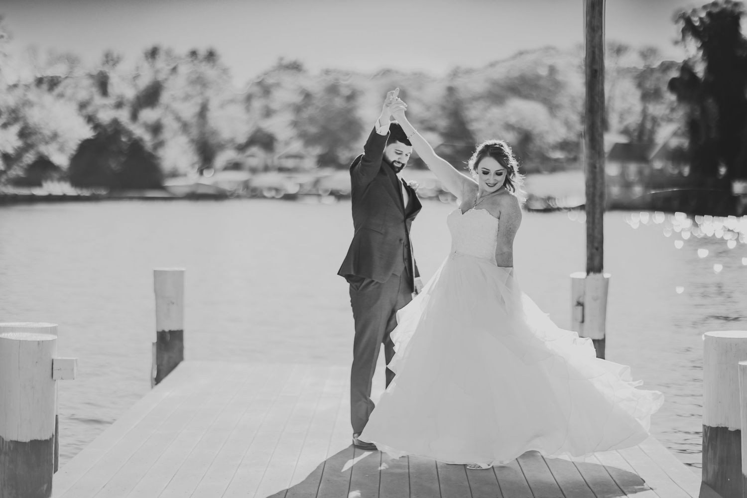 A black and white photo of newlyweds dancing on a dock by the water.