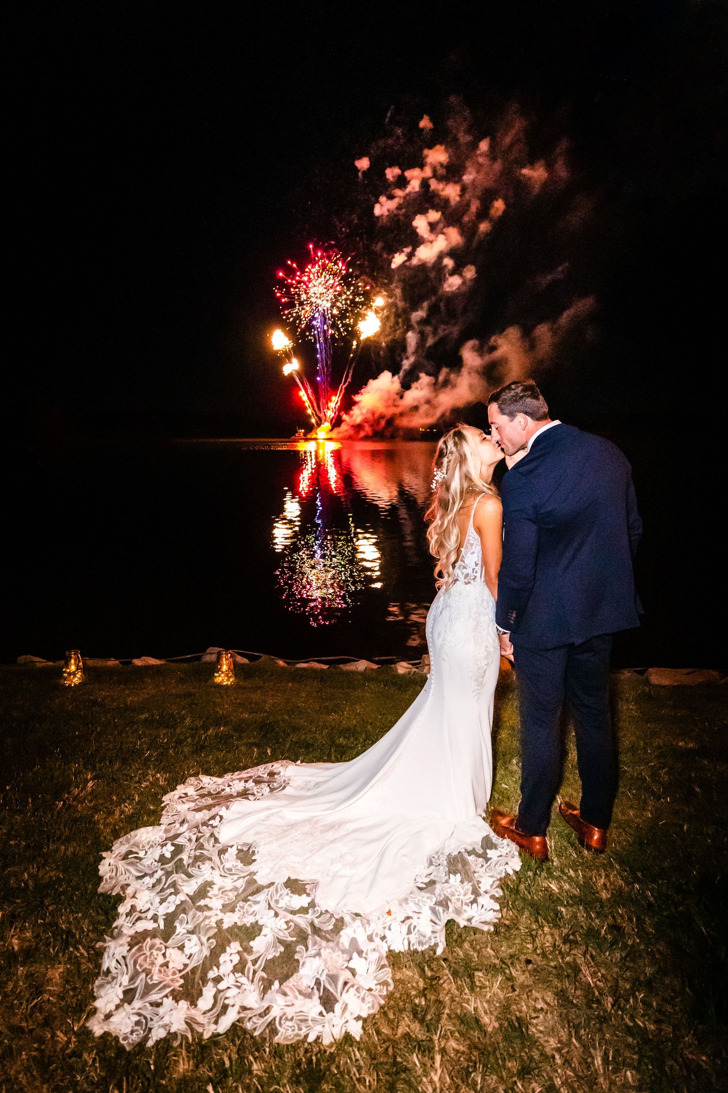 Wedding couple shares romantic moment under spectacular fireworks display beside a lake at night.