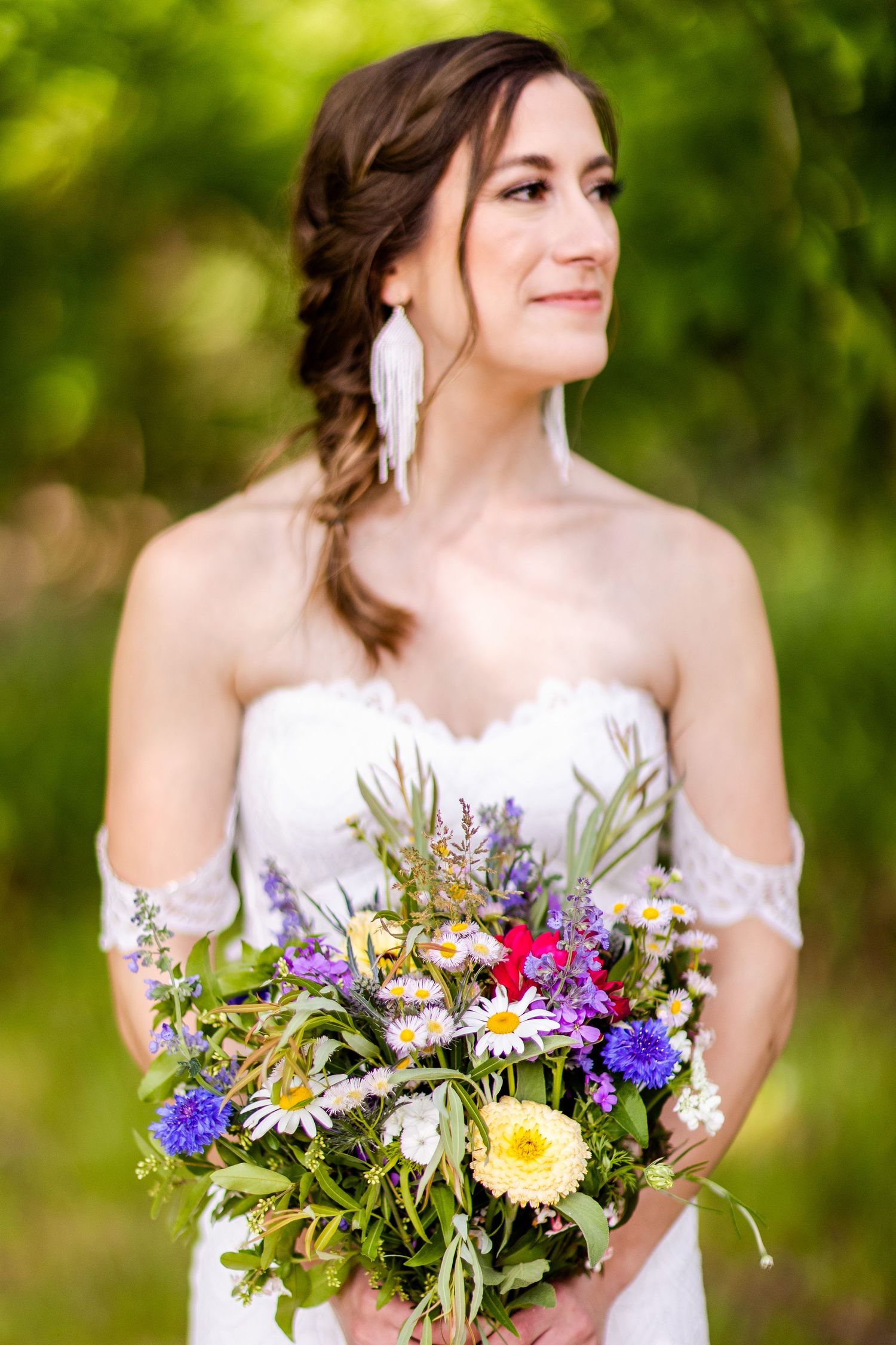 Bride in strapless dress holds vibrant wildflower bouquet with purple and yellow blooms in garden setting.