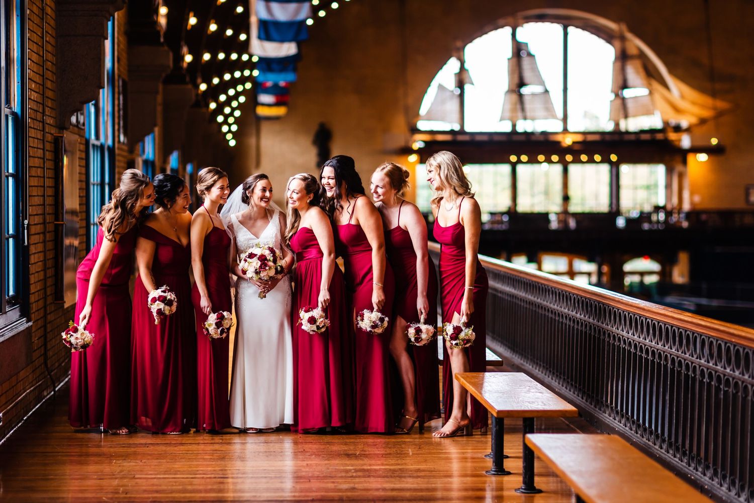 Wedding party in elegant red dresses posed in an ornate vintage venue with string lights.