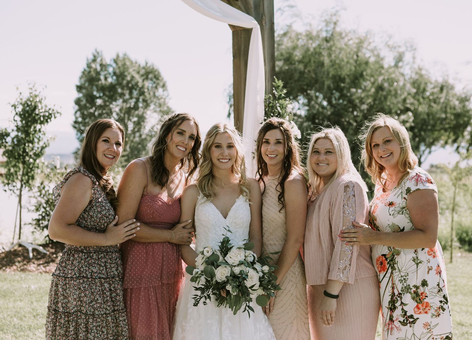Bride with bridesmaids in pastel dresses posing under decorated wedding arch.